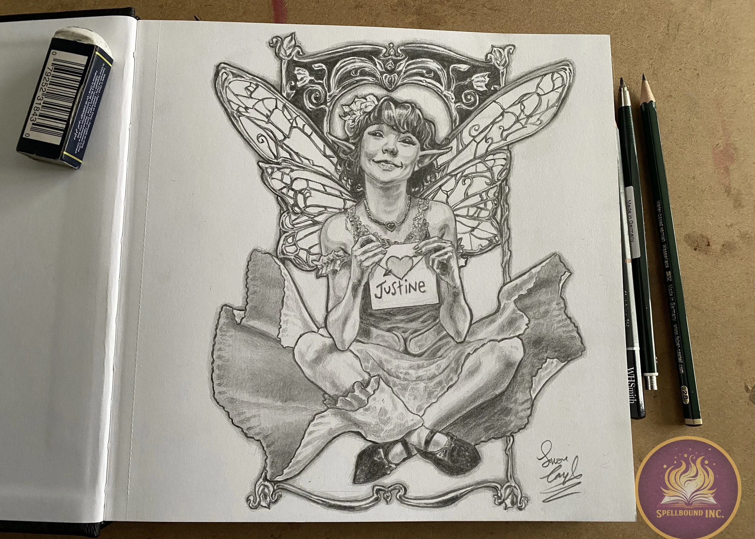 A detailed pencil drawing of a woman with fairy wings sitting cross-legged, holding a heart-shaped note with the name 'Justine'. She is smiling, wearing a dress and jewelry, with a decorative frame behind her, and is labeled with an artist's signatur
