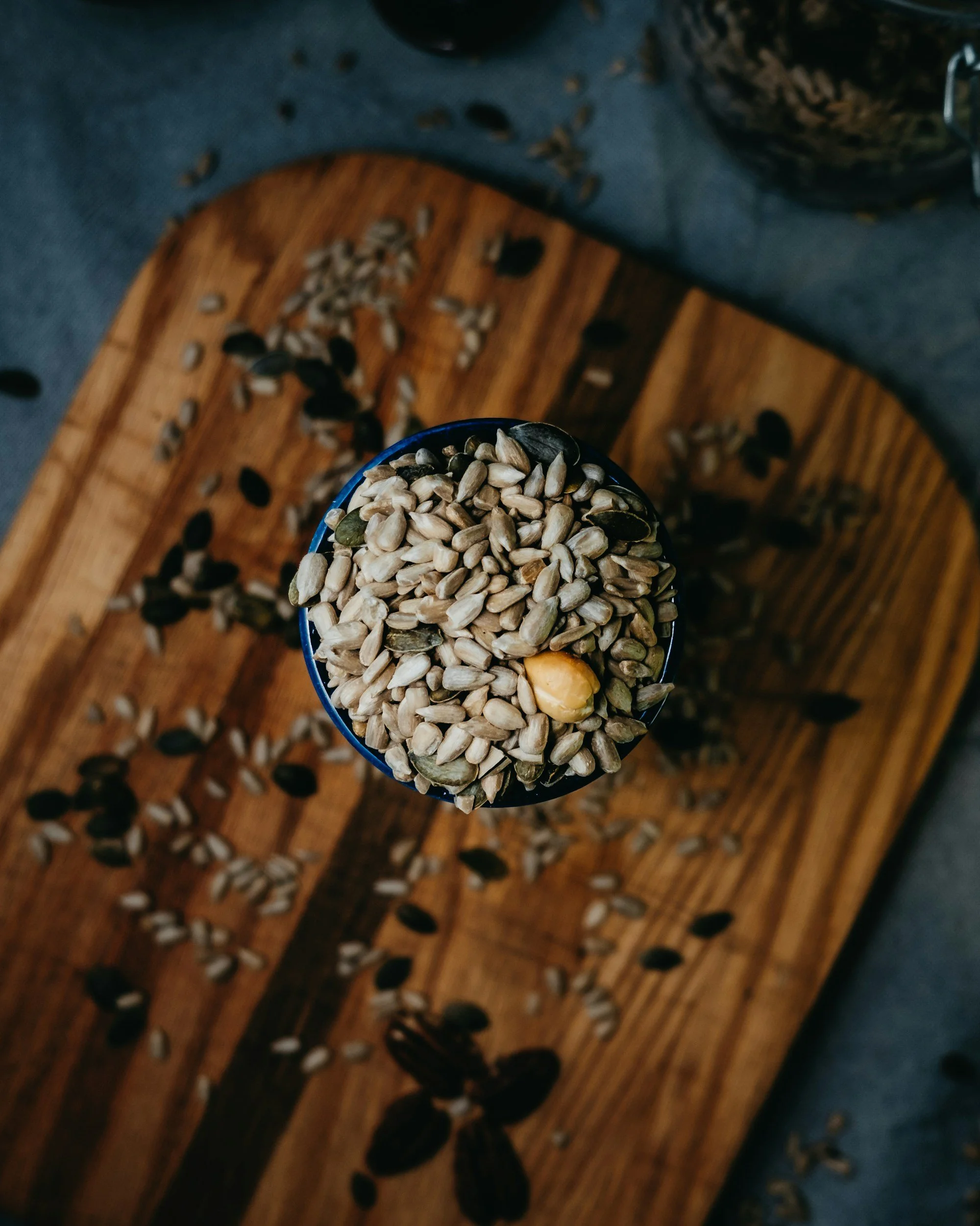 Close-up of a cup filled with mixed seeds and one yellow nut, placed on a wooden cutting board with scattered seeds and nuts around.