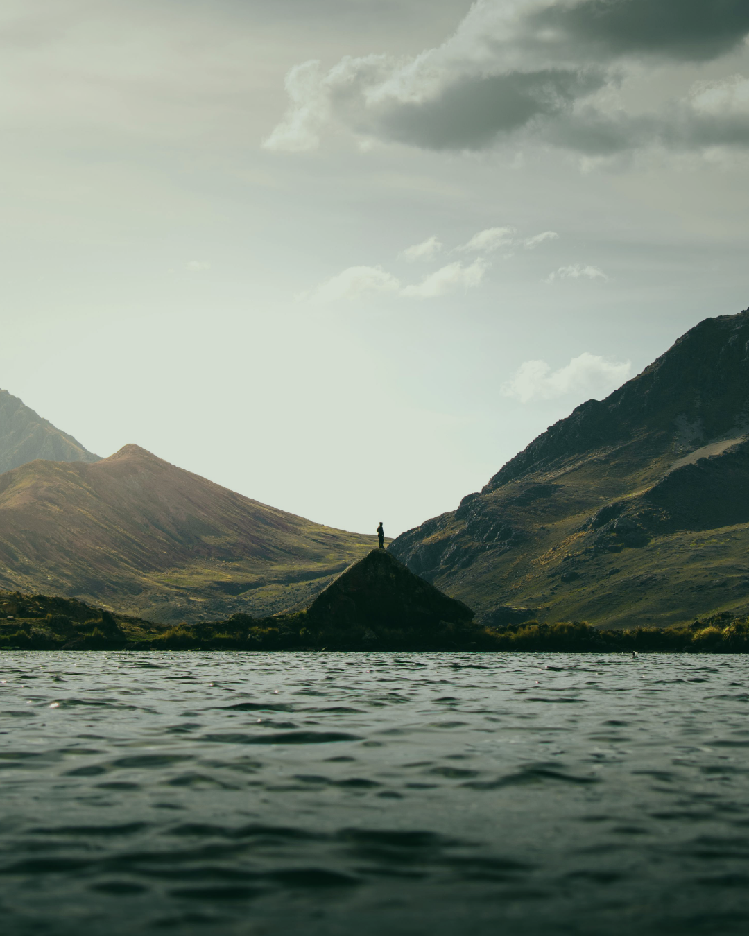 person standing on a small mountain on the other side of a lake