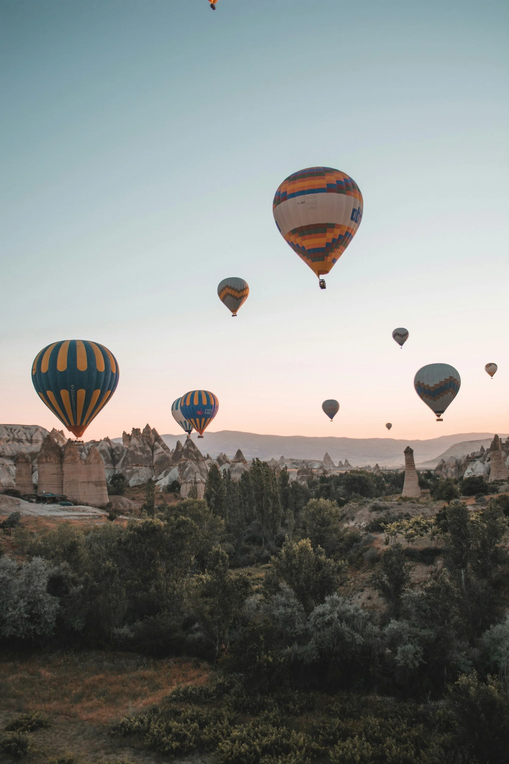 Multiple colorful hot air balloons floating over unique rock formations and green vegetation during sunrise or sunset.