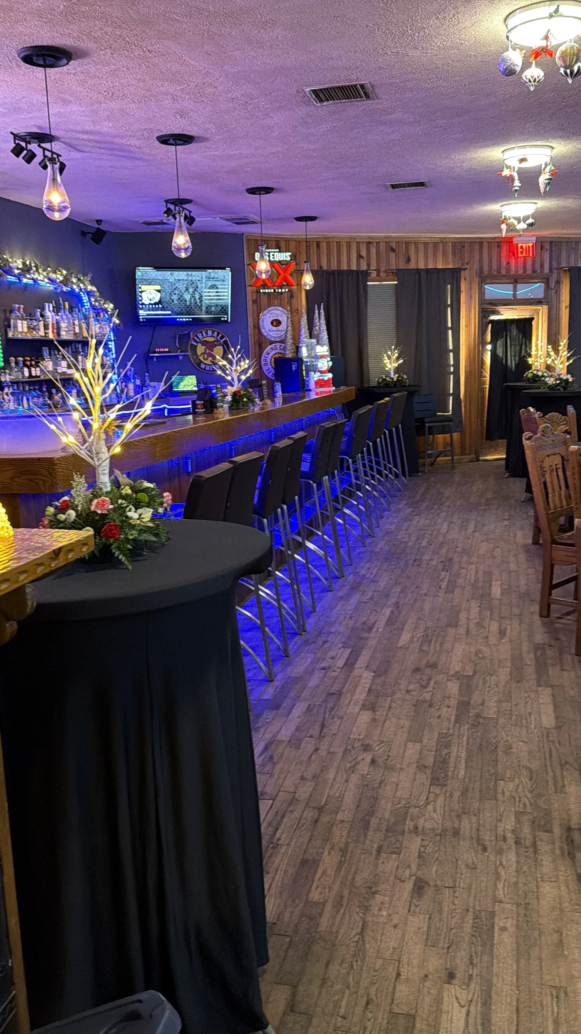 A cozy bar with wood-paneled walls, holiday decorations, and a row of high bar stools. There are Christmas centerpieces with lights and flowers on the tables and bar counter. The lighting is warm and festive, with hanging pendant lights and a decorated ceiling.