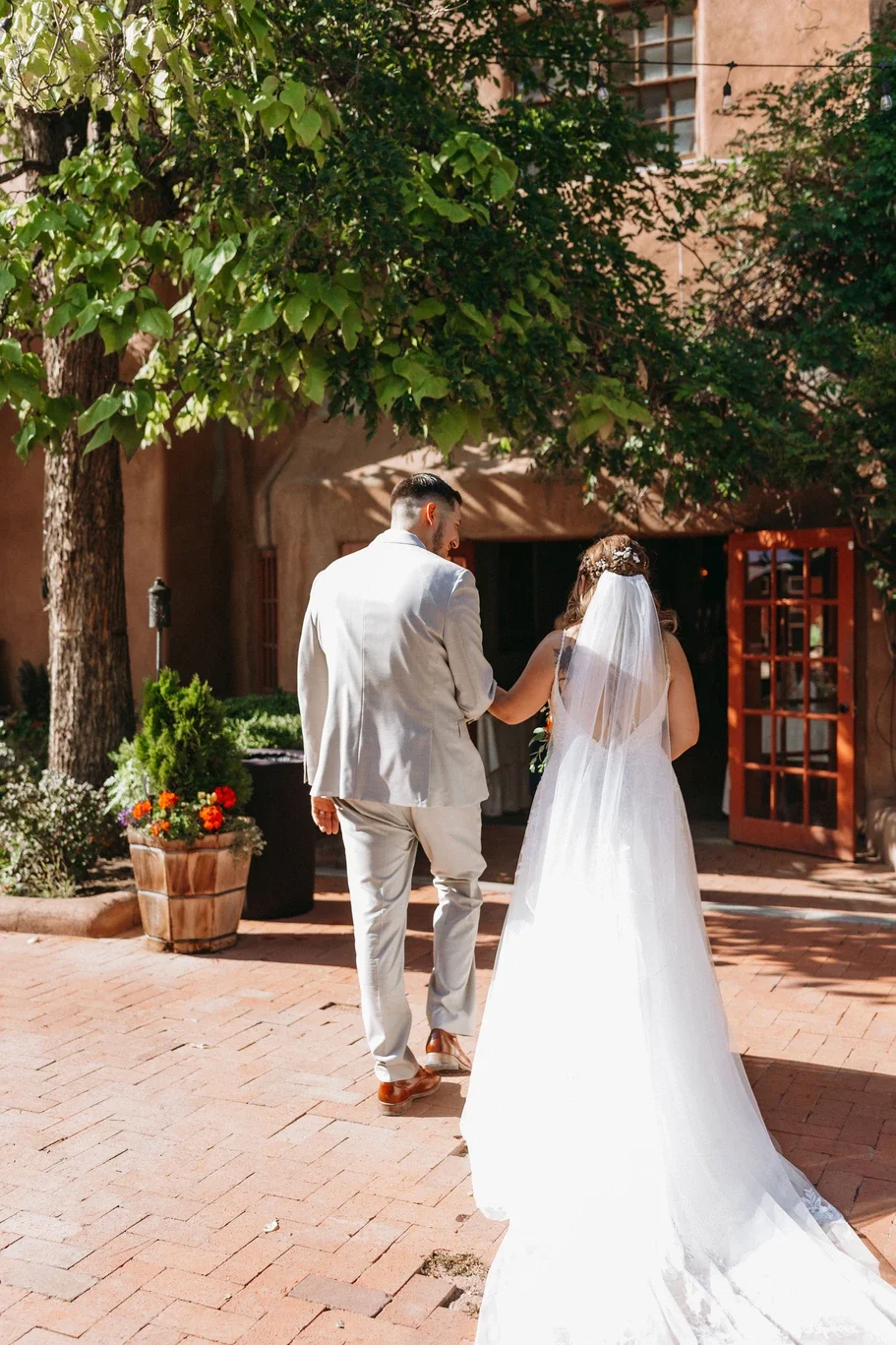 Wedding couple walking hand in hand outdoors, bride wearing a white wedding gown and veil, groom in a light-colored suit, brick-paved courtyard with trees and potted plants.