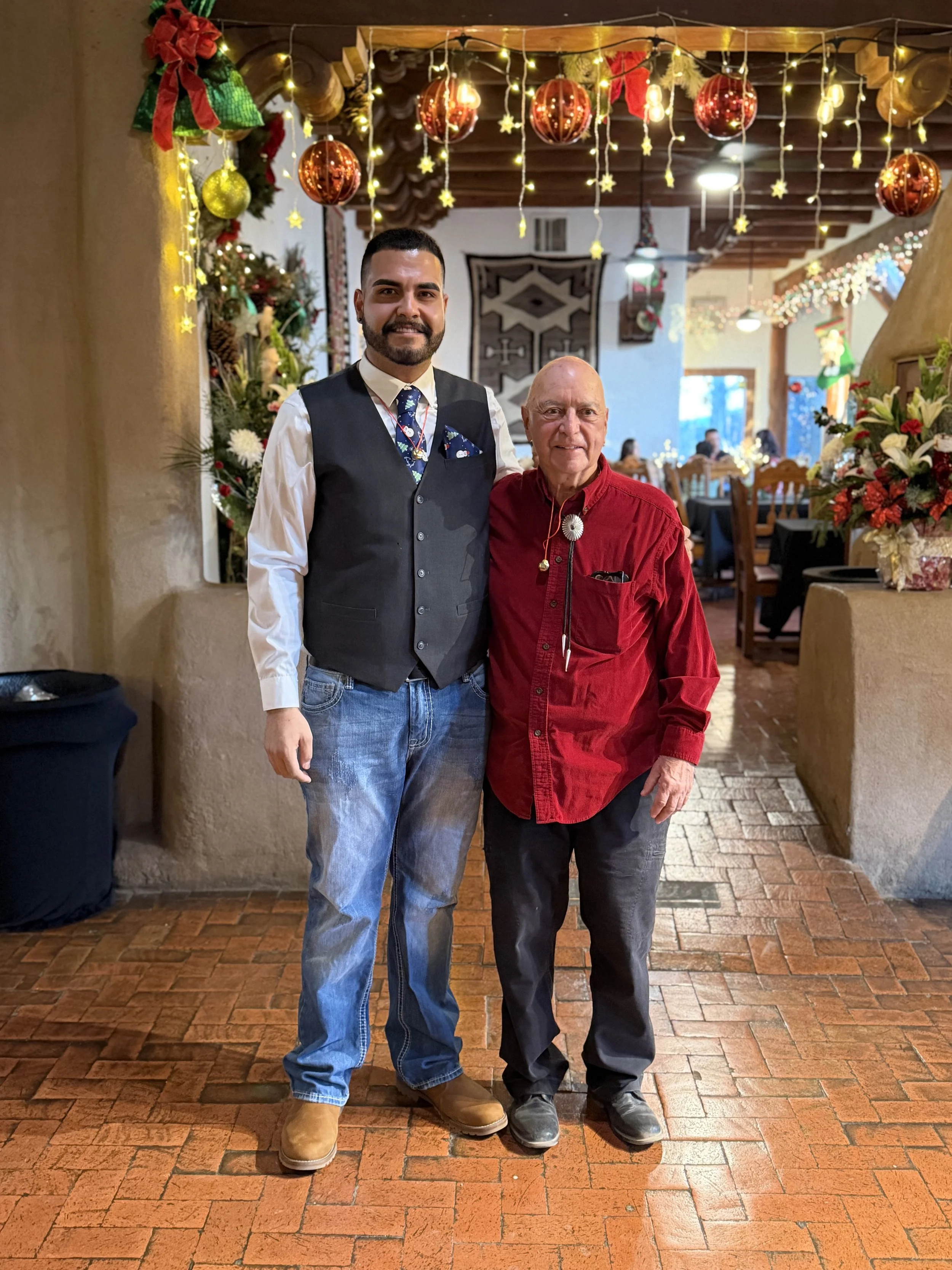 Two men standing close together and smiling in a decorated banquet hall for a Christmas celebration. The man on the left is tall with dark hair, a beard, wearing a black vest over a white shirt with a Christmas-themed tie, jeans, and tan boots. The man on the right is shorter, bald, wearing a red shirt with a bolo tie, dark pants, and black shoes. The background features Christmas decorations like hanging red and gold ornaments, string lights, Christmas trees, and poinsettias.