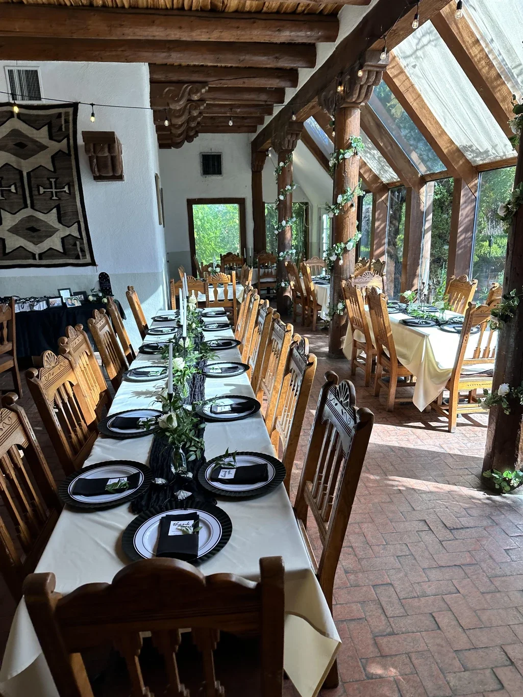 Long dining table set for a meal with black and white dishes, black napkins, white napkins, and white floral centerpieces, in a bright room with large windows, wooden beams, and string lights.