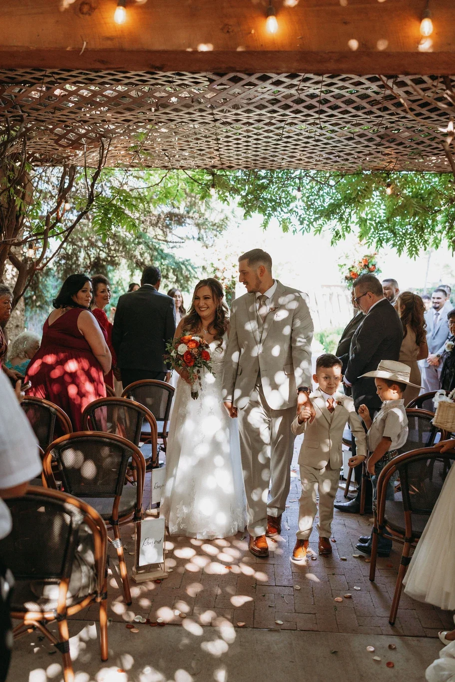 A bride and groom walking down the aisle at their outdoor wedding, surrounded by seated guests, children in formal attire, and decorated with trees and string lights.