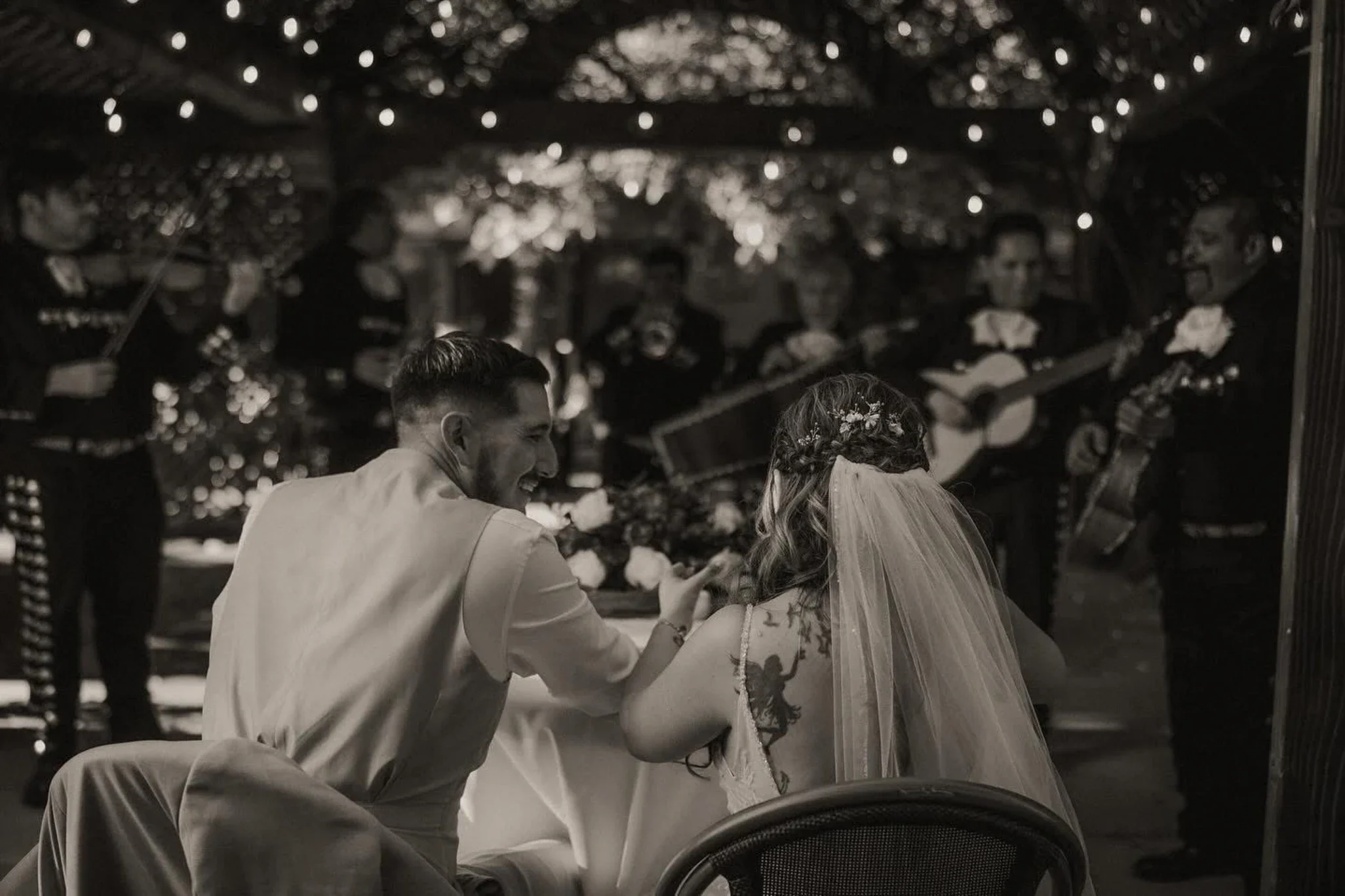 Black and white photo of a bride and groom sitting at a table, smiling; in the background, a mariachi band playing guitars and singing, performing under an outdoor, string-lit canopy.