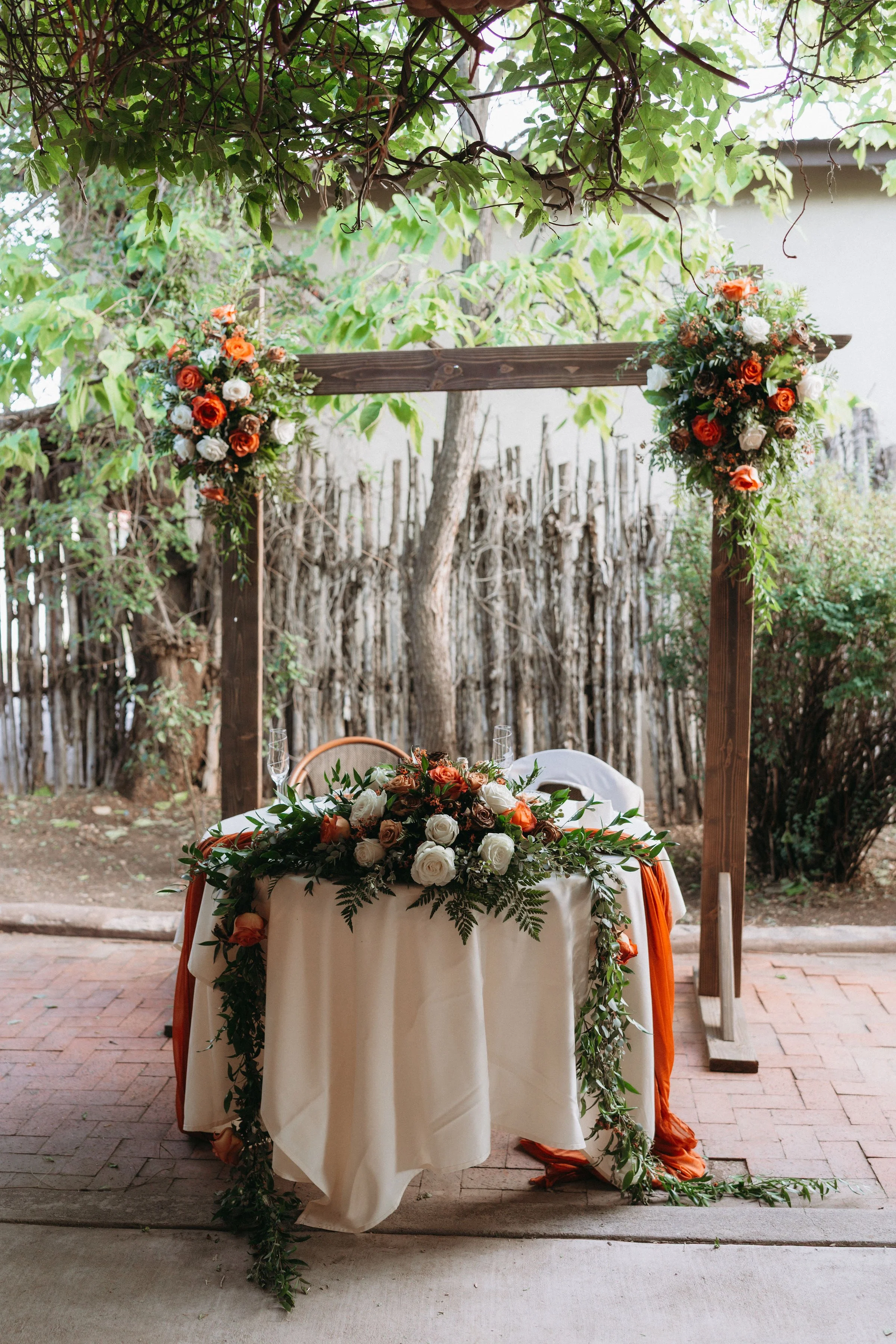 A table decorated with a floral arrangement under a wooden arch with floral bouquets, set outdoors on a brick patio, surrounded by trees and greenery.