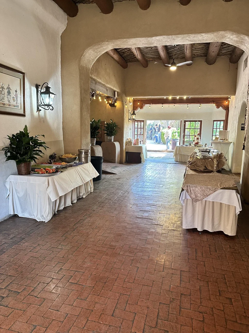 Indoor event space with tables covered in white tablecloths, decorated with plants, string lights, and food, leading to an outdoor patio area with people gathering outside.