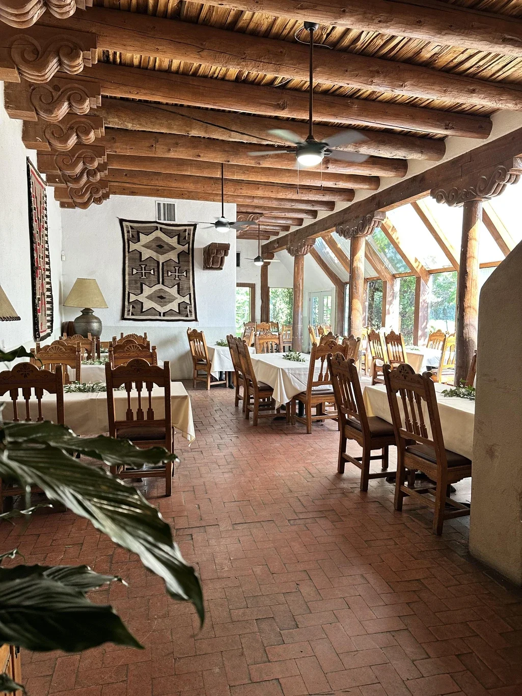 Interior of a rustic restaurant with wooden beams, chairs, and tables with white tablecloths, and decorative textiles on the wall.
