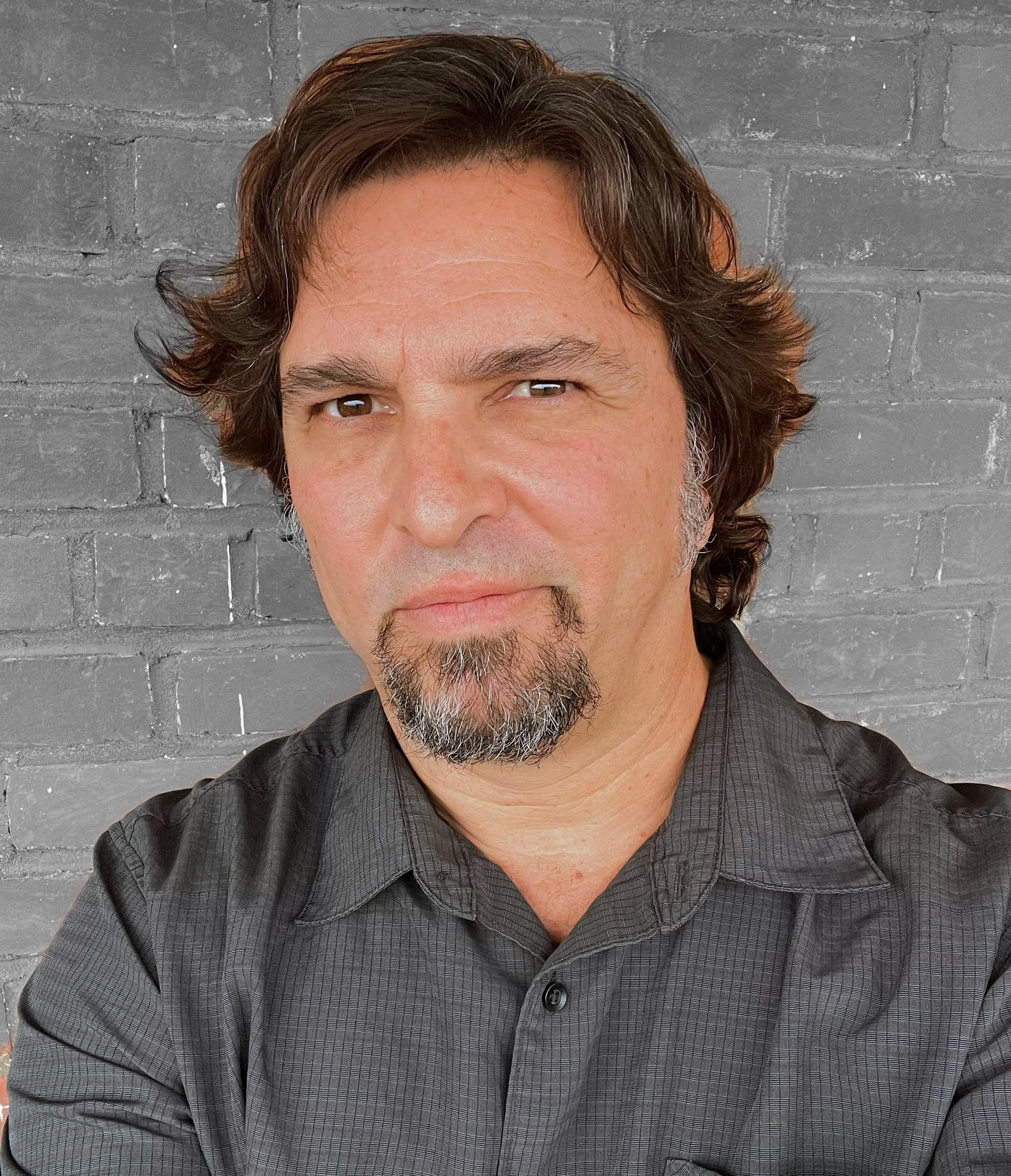 A man with shoulder-length brown hair, a goatee with some gray, wearing a dark gray button-up shirt, stands in front of a gray brick wall.