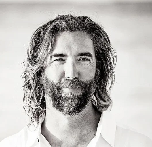 Black and white portrait of a middle-aged man with long wavy hair and a beard, smiling slightly, wearing a white shirt.