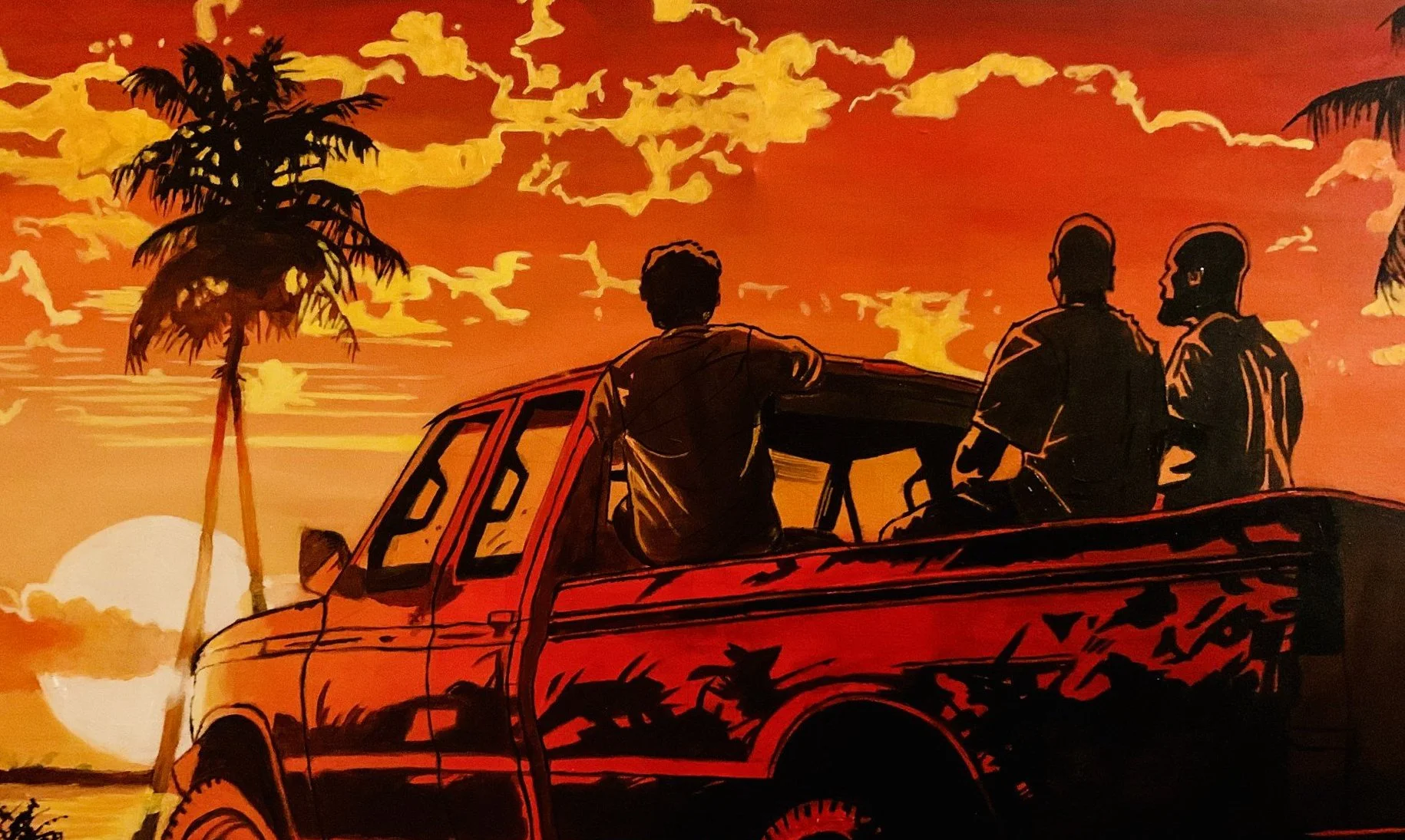 Silhouette of three men in the back of a pickup truck watching a Caribbean island sunset with palm trees and clouds in the sky.