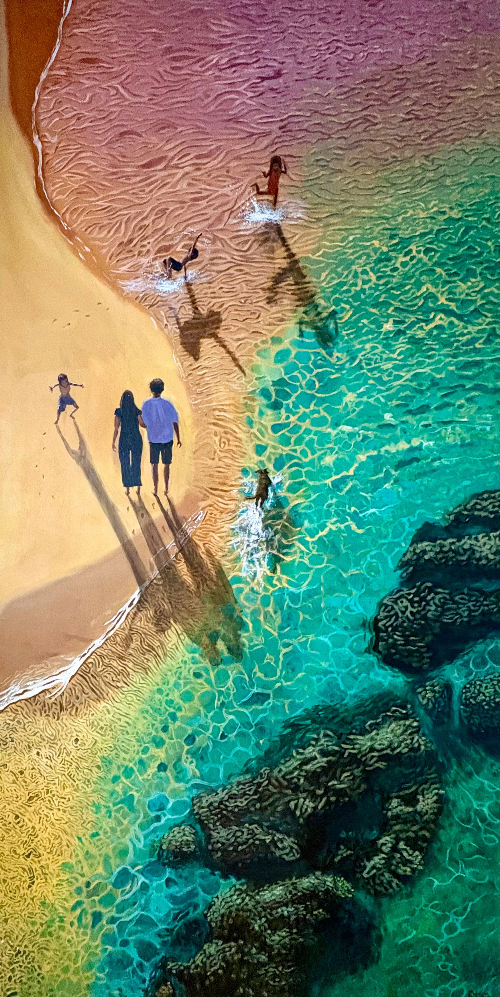 Painting of an aerial view of a beach with golden sand, where children are swimming and playing in the water. An adult couple walks along the shoreline, casting long shadows, while others are in the water near rocks.