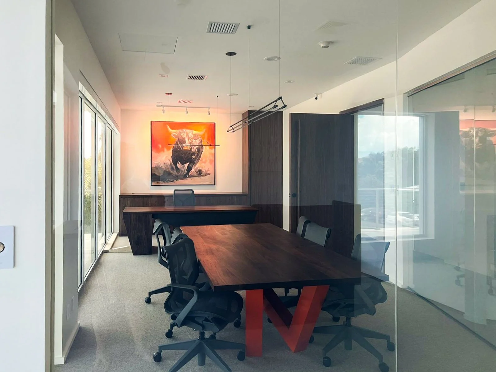 Modern conference room with a wooden table, six black office chairs, a tile floor, windows on the left, a colorful art piece of a bull on the wall, and a glass wall on the right.