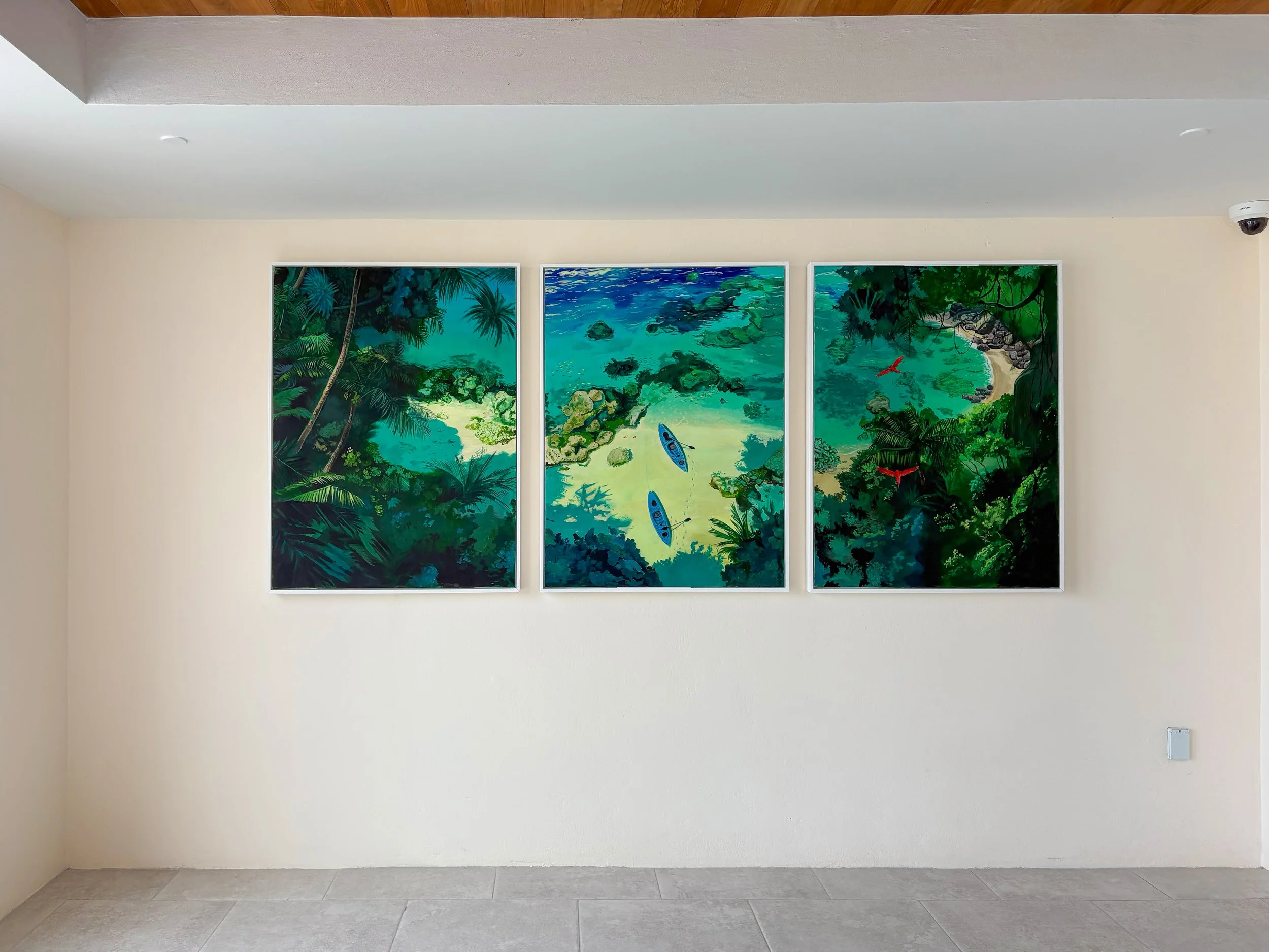 A triptych painting of a tropical jungle scene with a beach, ocean, and palm trees, mounted on a white wall in a modern interior.