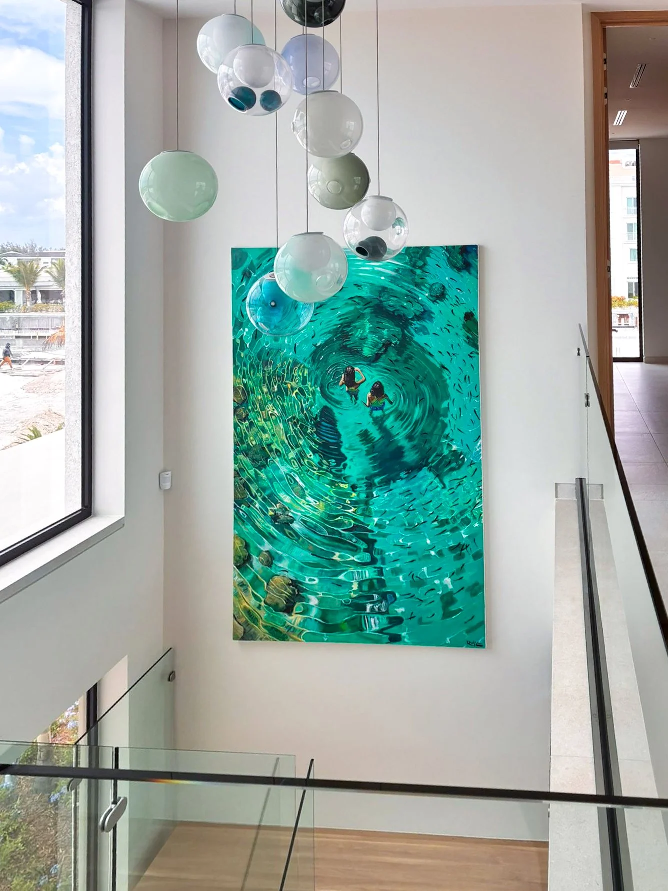 Interior view of a modern building with a large hanging light fixture, a painting of two people swimming in a pool, and a staircase with glass and metal railings.