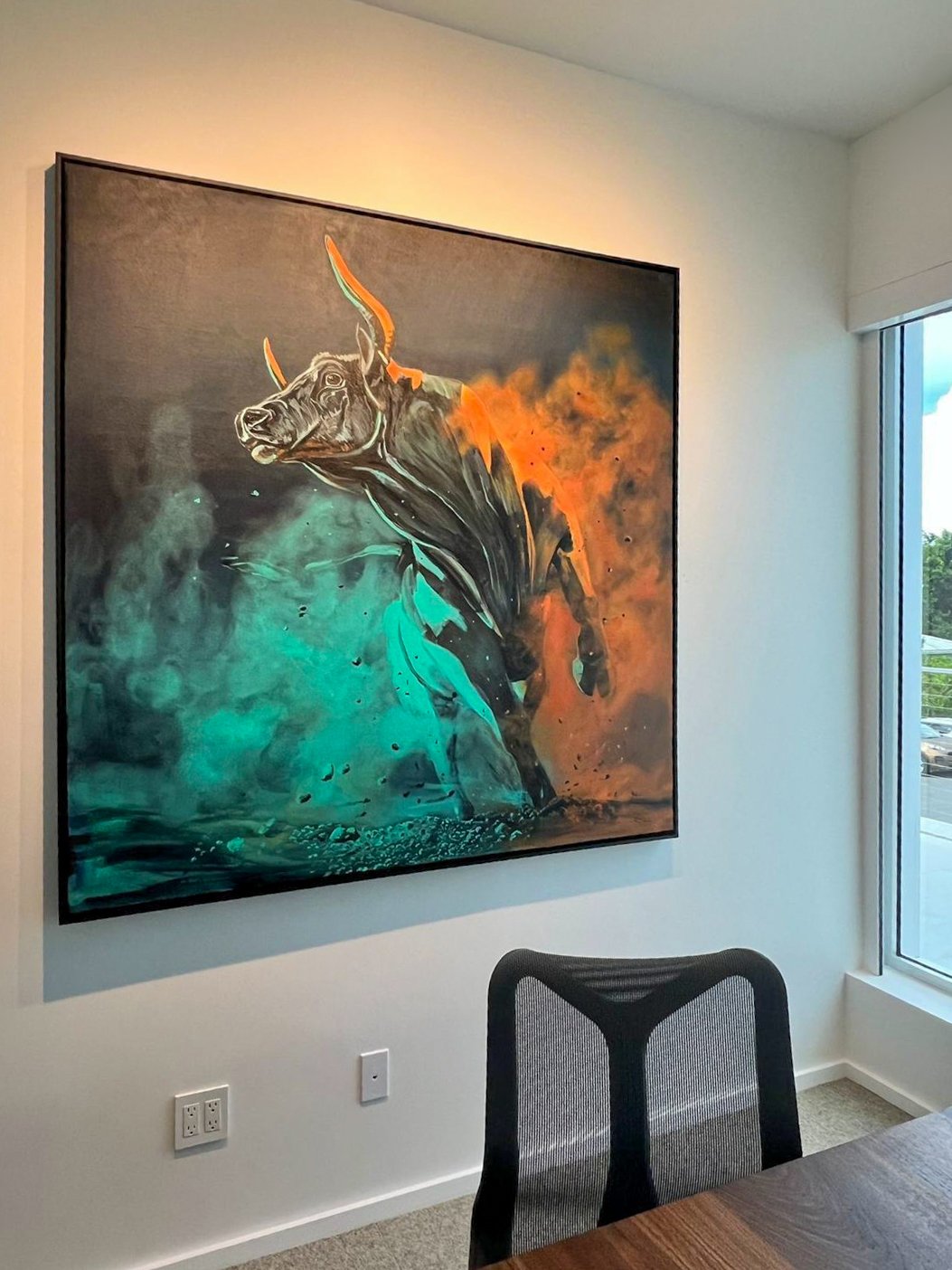 Colorful modern painting of a bull with dynamic brushstrokes in teal and orange hues hung on a white room wall, near a window.
