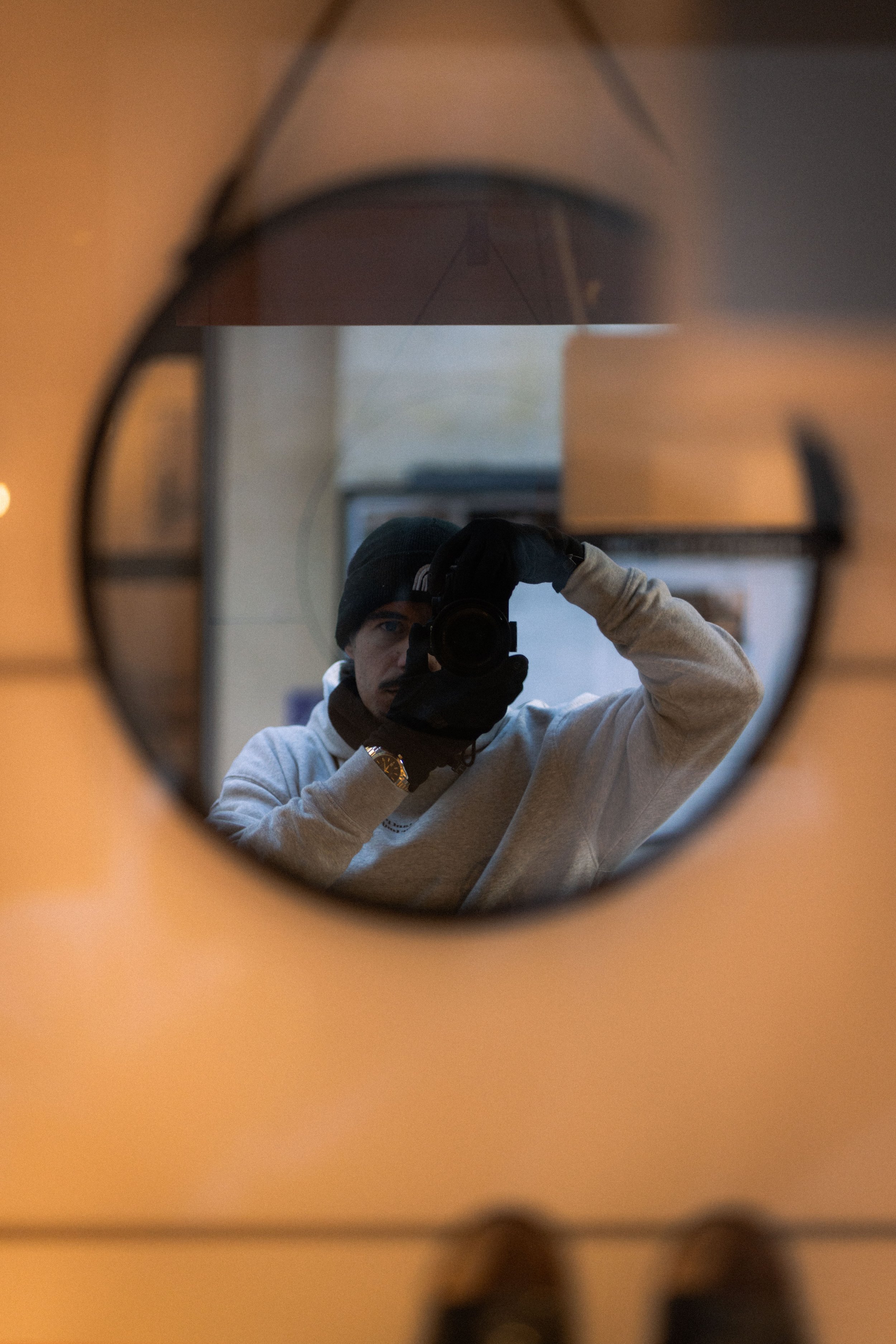 Un homme portant un bonnet noir, un sweatshirt gris, et des gants noirs, se prend en photo dans un miroir, tenant un appareil photo.