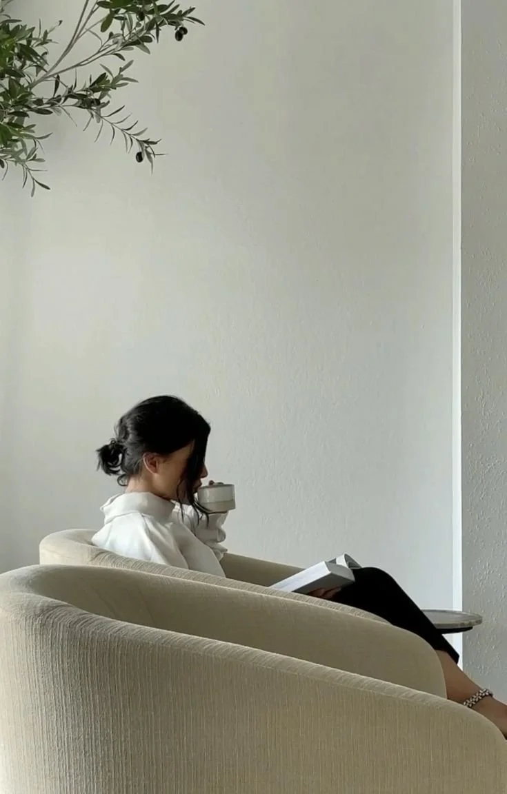 A woman sitting on a beige sofa, reading a book and drinking from a mug, with an olive tree branch visible in the upper left corner of the image.