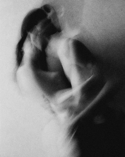 Black and white photo of a nude woman with long hair covering her face, sitting on the floor, hugging her knees.