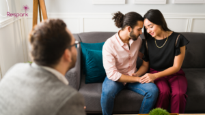 A woman and a man sitting close together on a gray couch, smiling and holding hands, while a therapist or counselor listens in front of them during a therapy session.