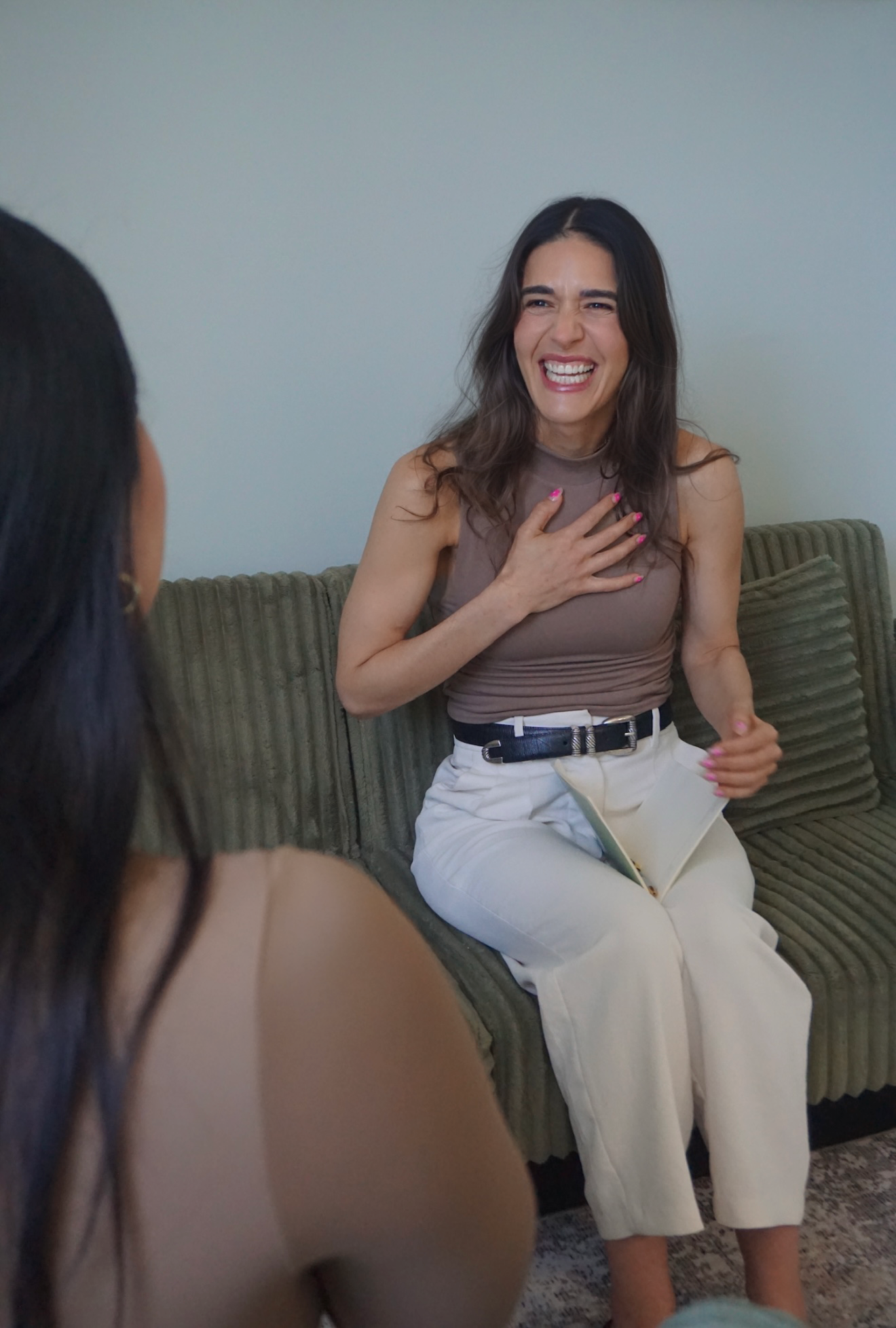 A woman with dark hair, smiling and touching her chest, sitting on a green striped couch, engaging with another person whose arm and part of their hair are visible in the foreground. The woman is dressed in a sleeveless top and white pants, holding an open book.