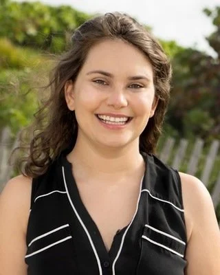 A young woman with brown curly hair smiling outdoors with green trees and a white fence in the background.