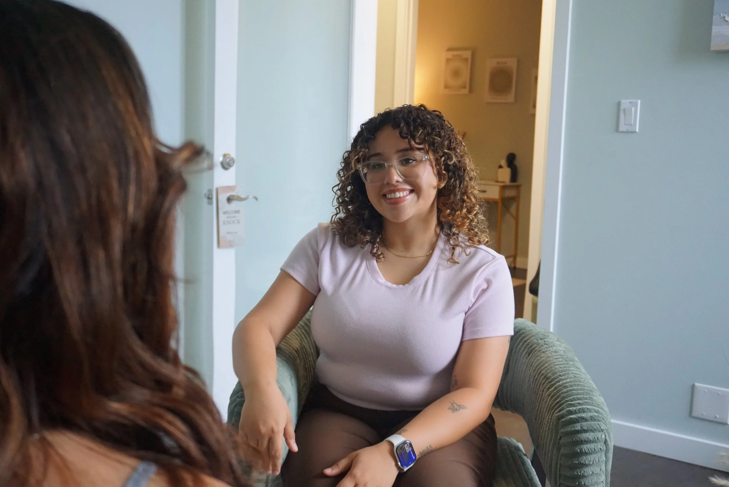 A woman with curly hair and glasses sitting on a green armchair, smiling and talking to another person in a house.
