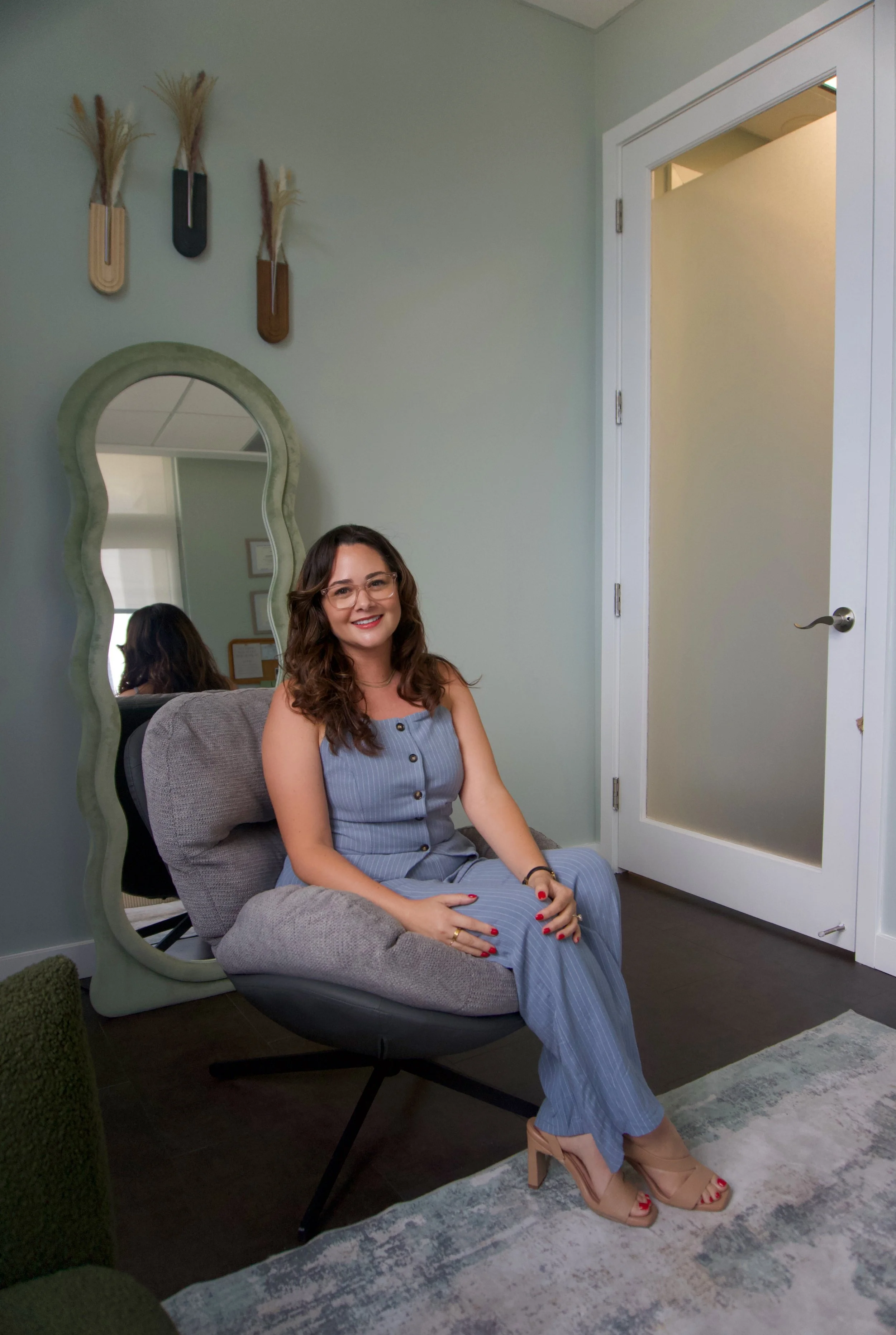 A woman with wavy brown hair, glasses, and a blue striped jumpsuit sitting on a gray cushioned chair with her legs crossed, smiling, in a room with light green walls and a decorative mirror above her. She has red nail polish, beige high heels, and a black bracelet. A large green fuzzy ottoman and a patterned rug are also visible.