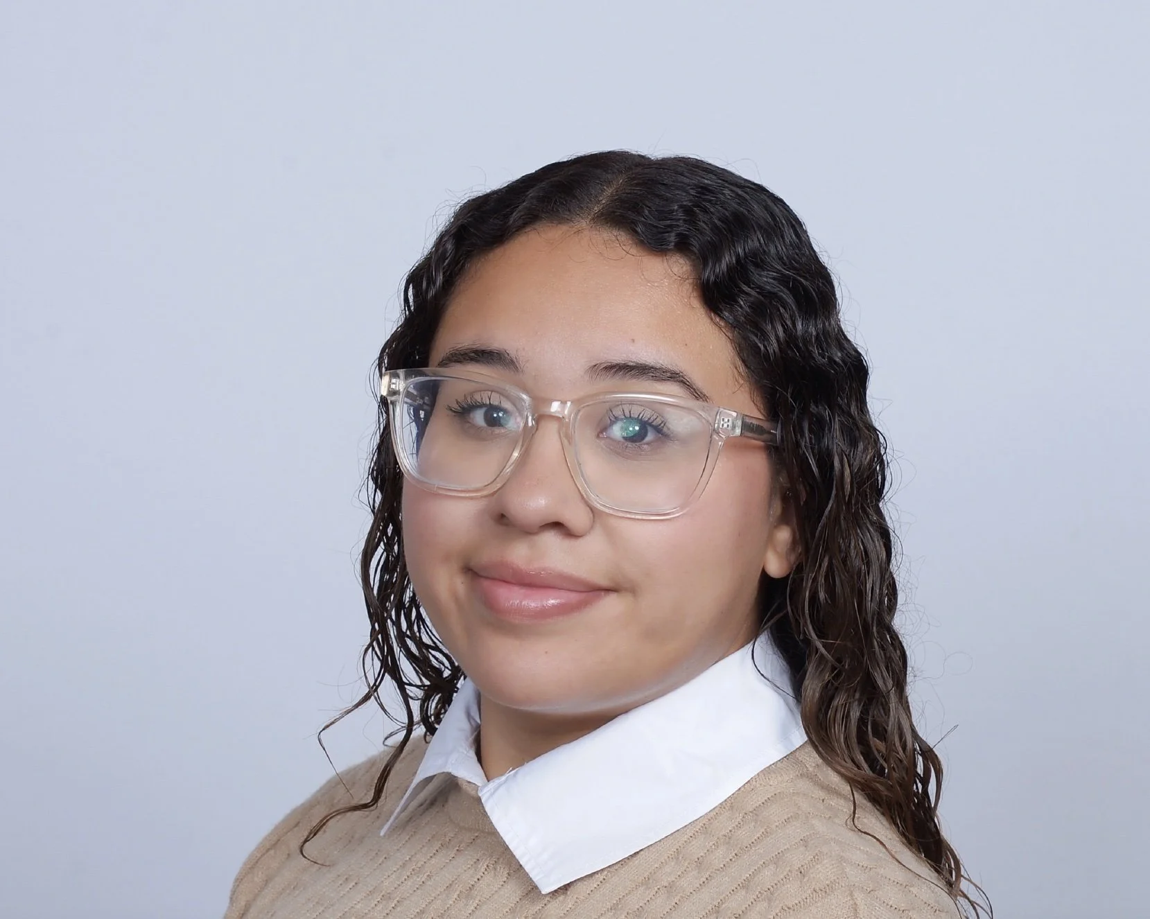 Portrait of a young woman with curly hair wearing glasses, a white collared shirt, and a beige sweater, smiling softly against a light gray background.