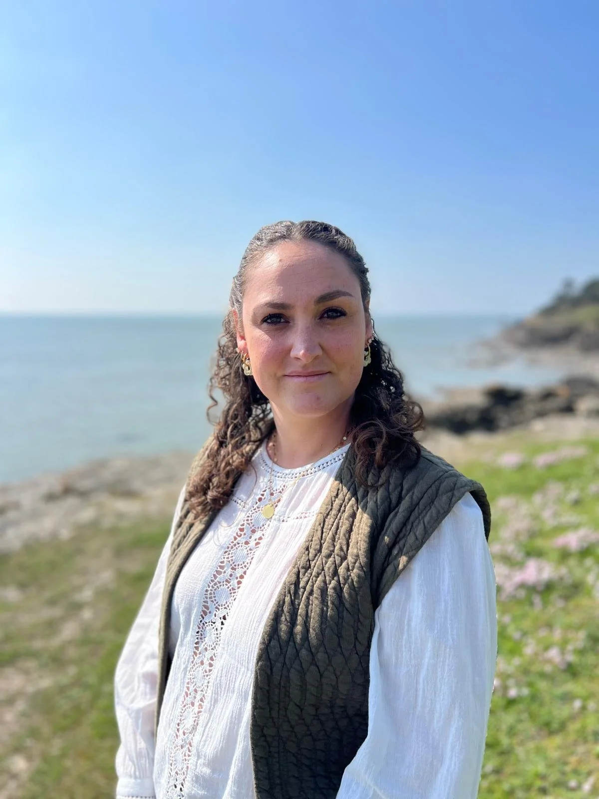 Femme avec des boucles d'oreilles en or, portant une chemise blanche avec des broderies, un gilet vert en tissu matelassé, posant à côté de la mer avec un ciel bleu clair. Doula
