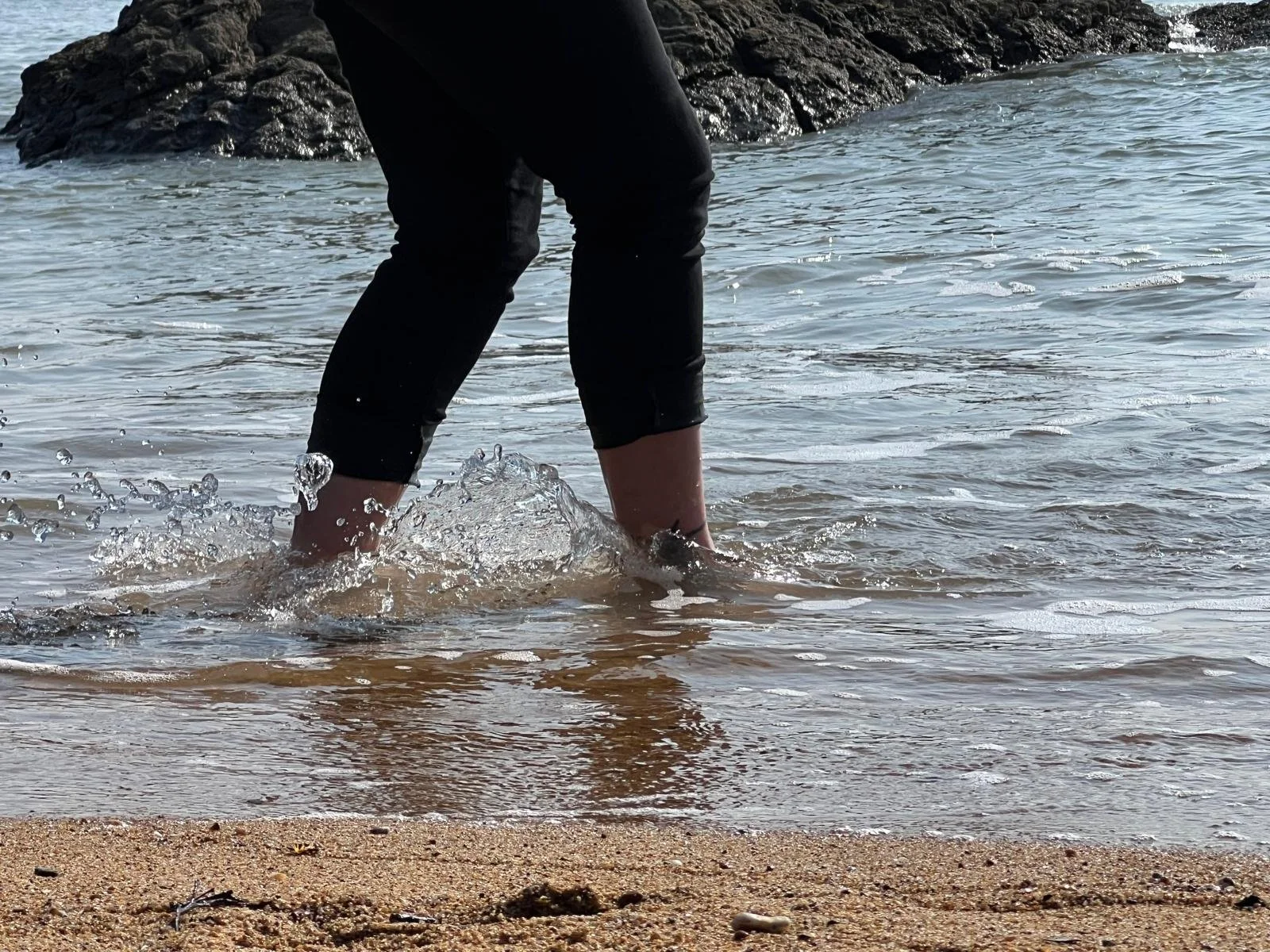 Une personne portant un pantalon noir marche dans la mer, avec des vagues l'entourant, devant une roche, regardant vers la mer. Doula