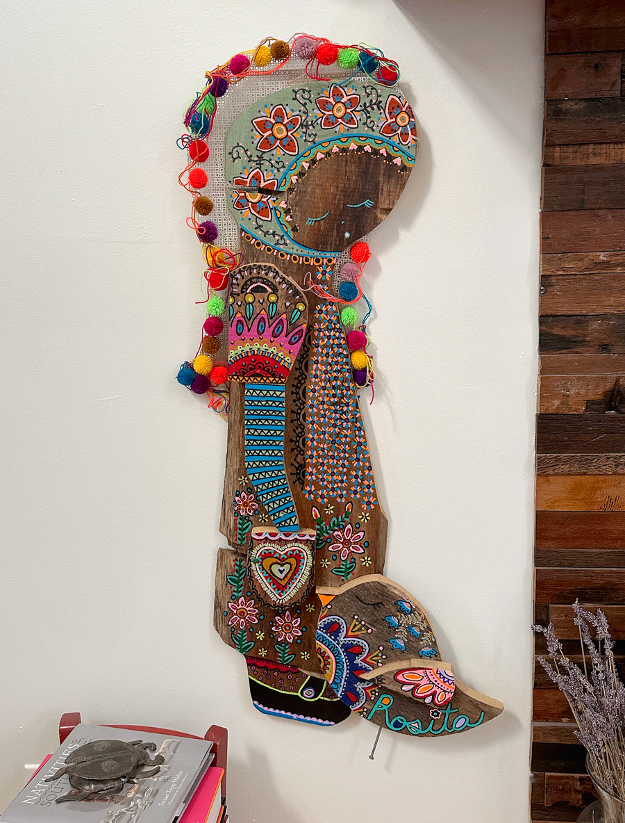 Decorative wooden wall hanging featuring a stylized portrait of a girl with closed eyes and plants. The artwork is colorful and detailed, with a multicolored yarn border decorated with pom-poms.