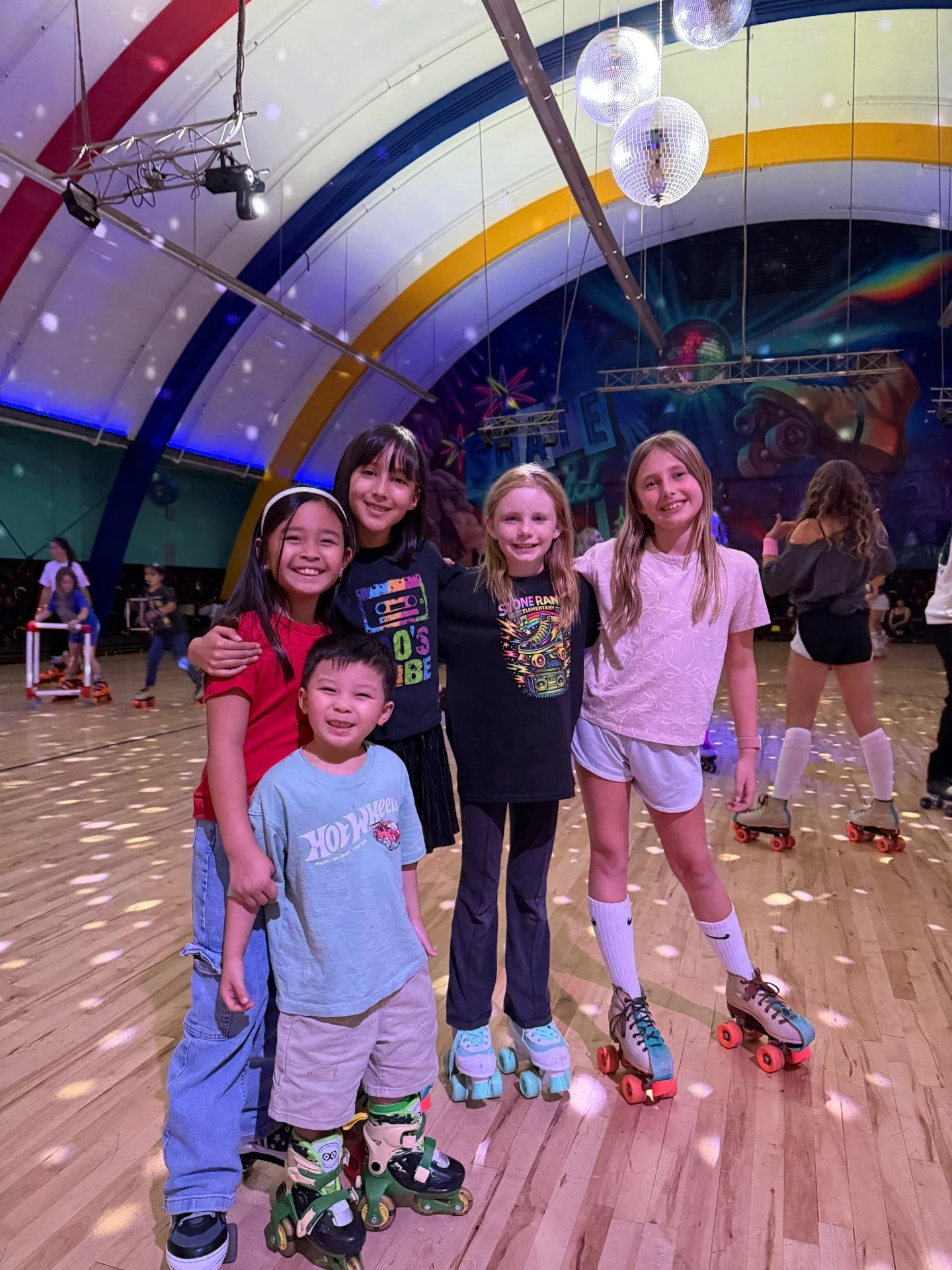 Five children with roller skates standing together in a roller skating rink, smiling at the camera, with disco balls and colorful lights overhead, and other skaters in the background.
