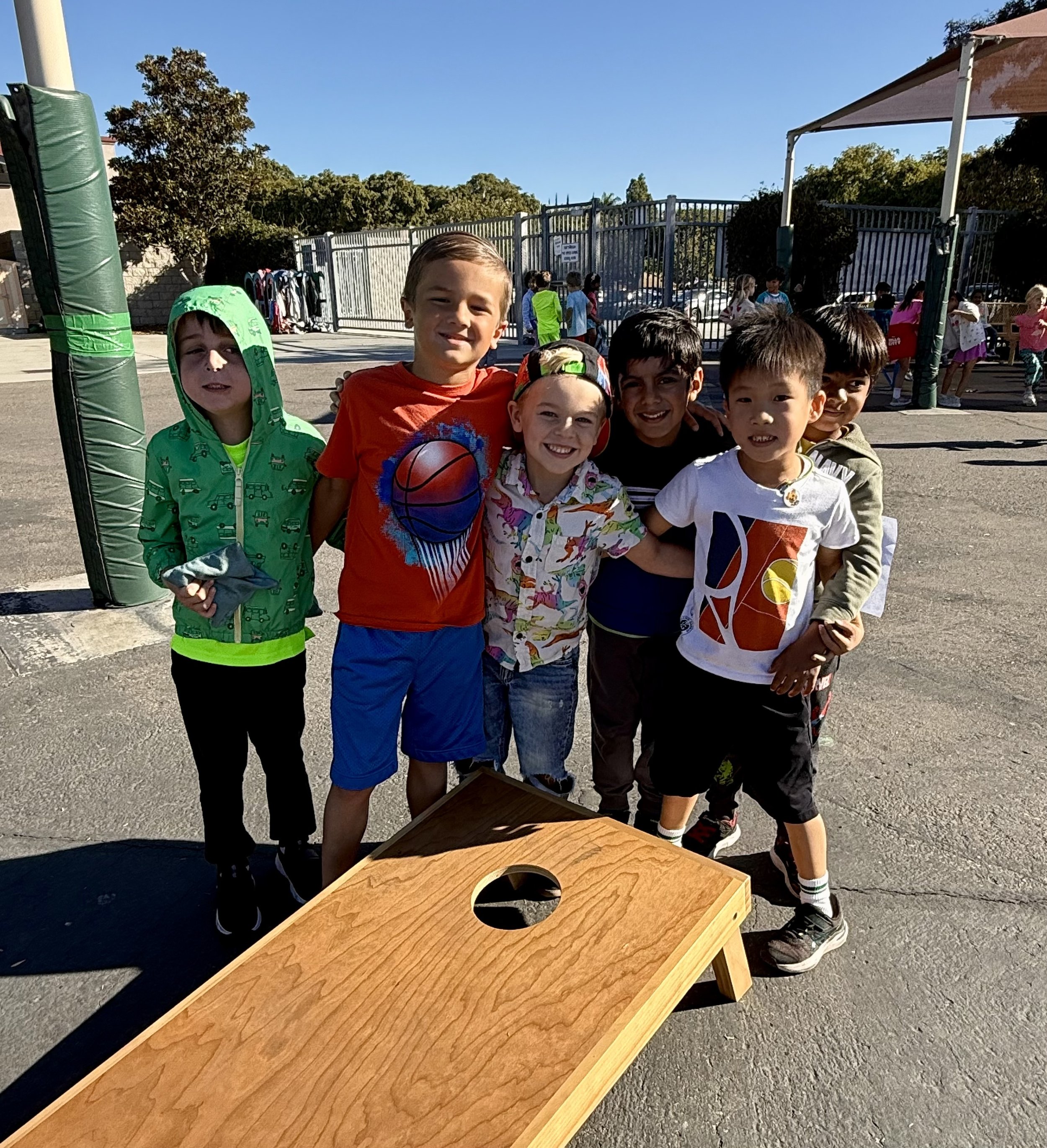 Group of children standing outdoors near a cornhole game, smiling at the camera, with a fence, trees, and other children playing in the background on a sunny day.