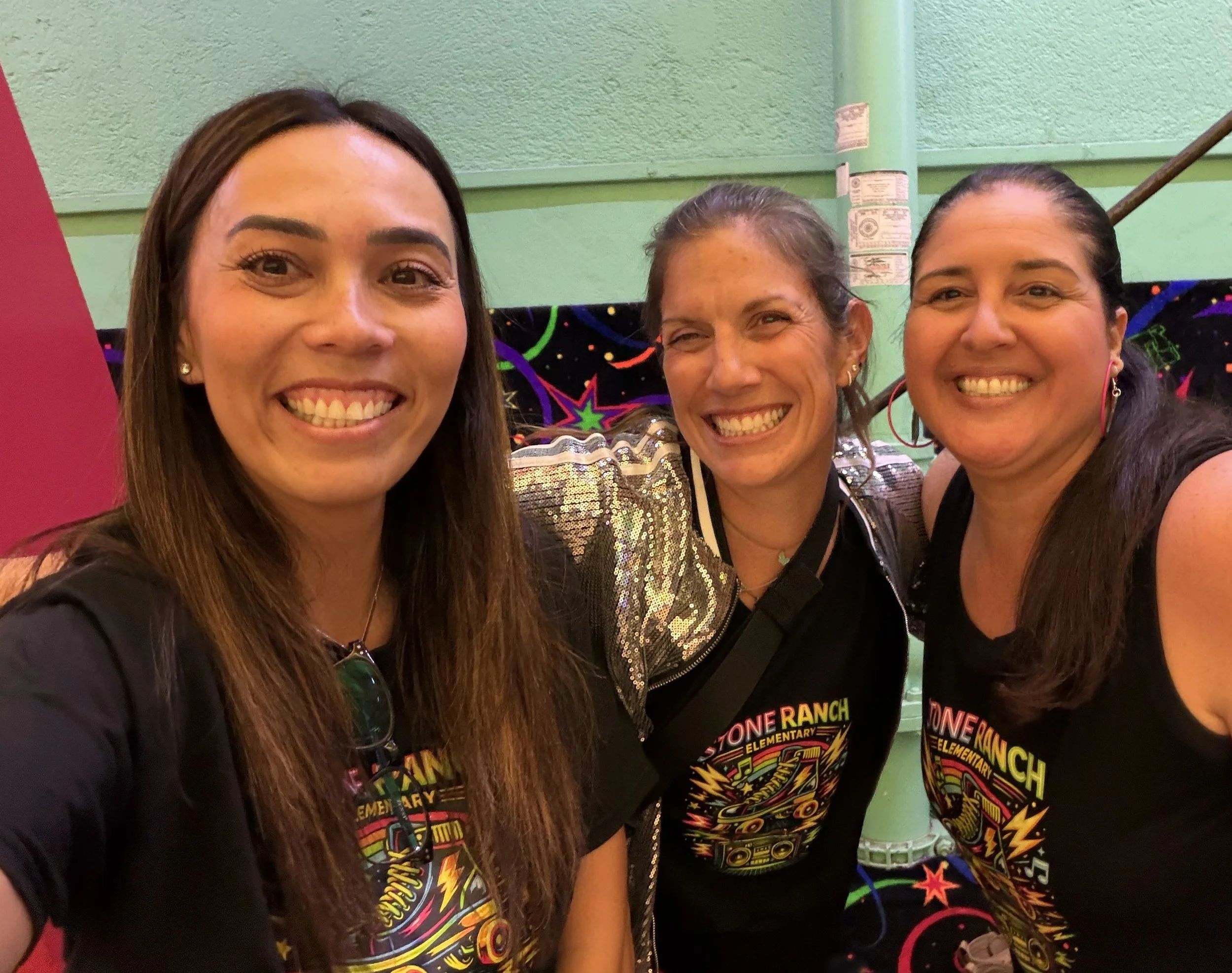 Three women smiling, close-up selfie, wearing black T-shirts with colorful graphics related to Stone Ranch Elementary School, with a green wall and a colorful backdrop in the background.