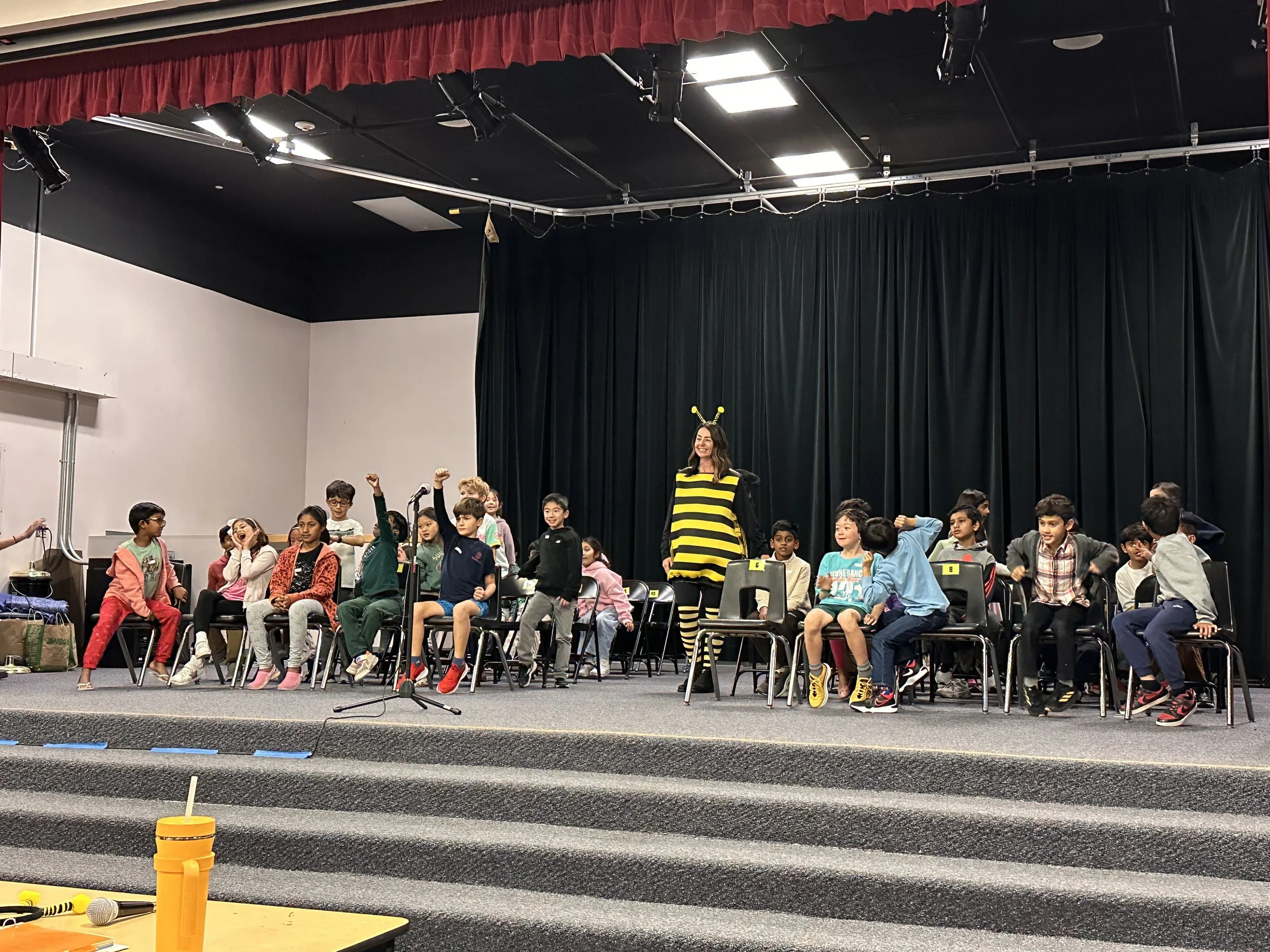 Children and a woman dressed as a bumblebee on stage at a school event, with children sitting on chairs in front and a black curtain backdrop.
