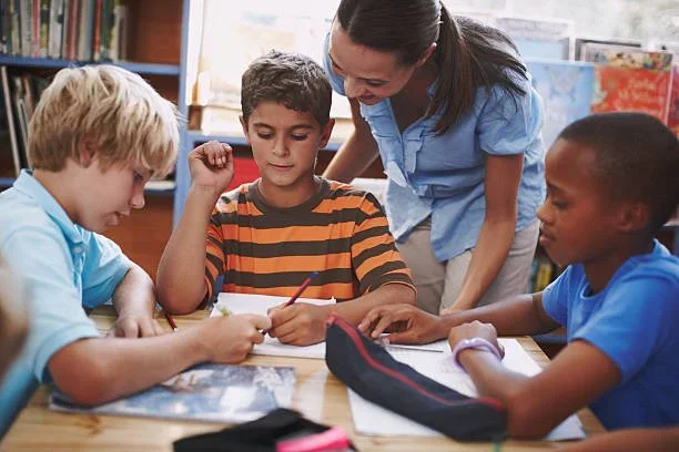 A teacher helps three children with schoolwork at a table in a classroom.