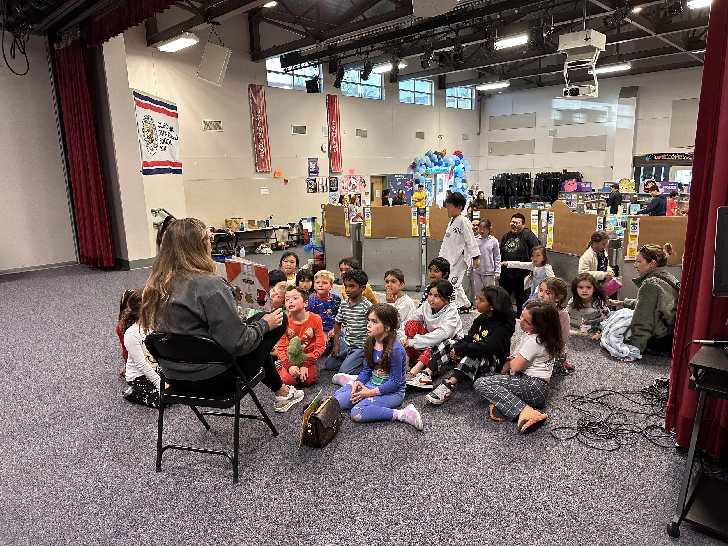 Youth group gathered on the floor of an auditorium, listening to a woman reading a book. The room has banners, balloons near the entrance, and a stage with red curtains, suggesting a school or community event.