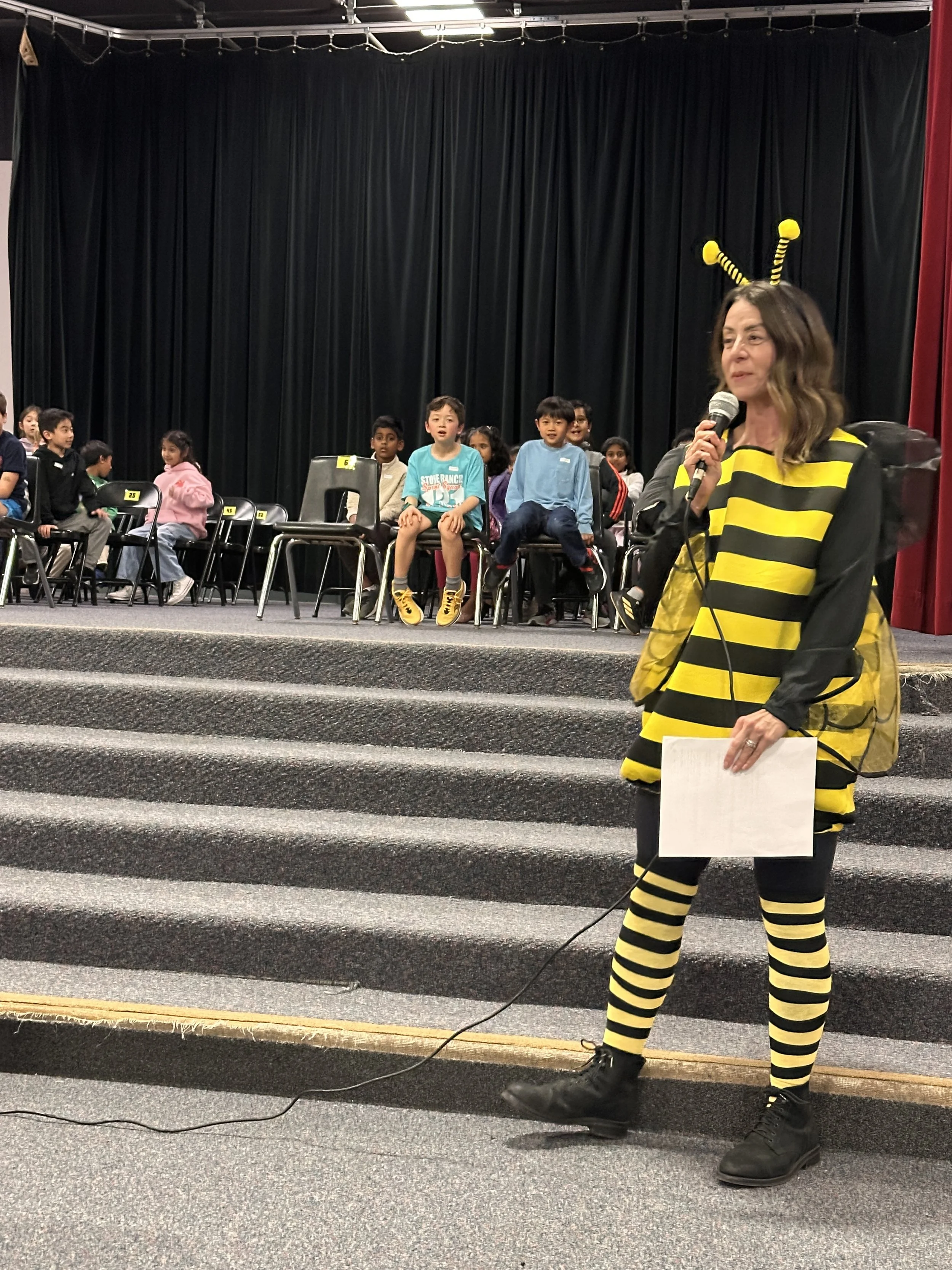 A woman dressed as a bee holding a microphone and a sign, standing on a stage with gray carpet and stairs. Behind her, children are seated in chairs in front of black curtains.