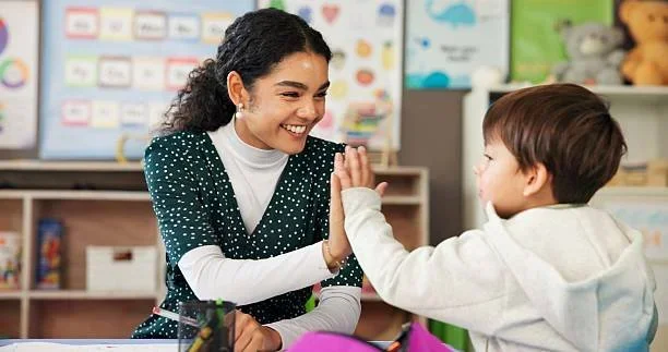 A teacher smiling and giving a high five to a young student in a classroom.