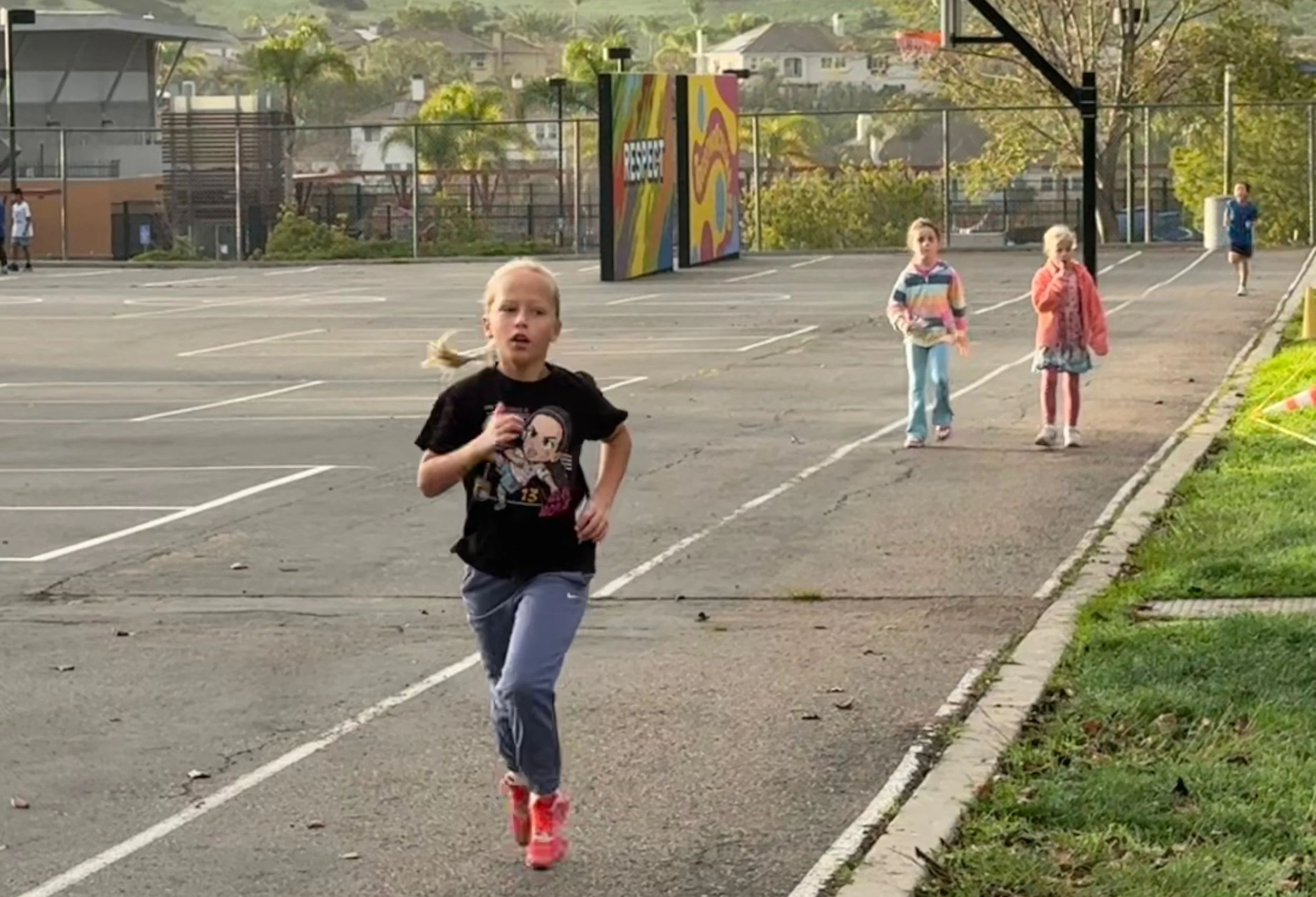 Young girl running on an outdoor sports court with other children in the background, some walking and some running, surrounded by a fence and residential houses with trees in the distance.