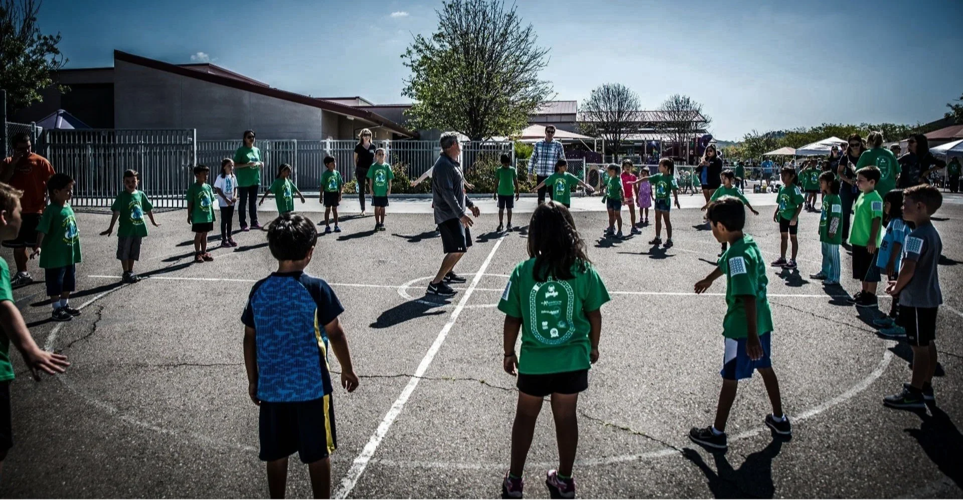 Children participating in an outdoor group activity on a school playground, standing in two lines facing each other, with adults supervising, under clear skies with trees and school buildings in the background.