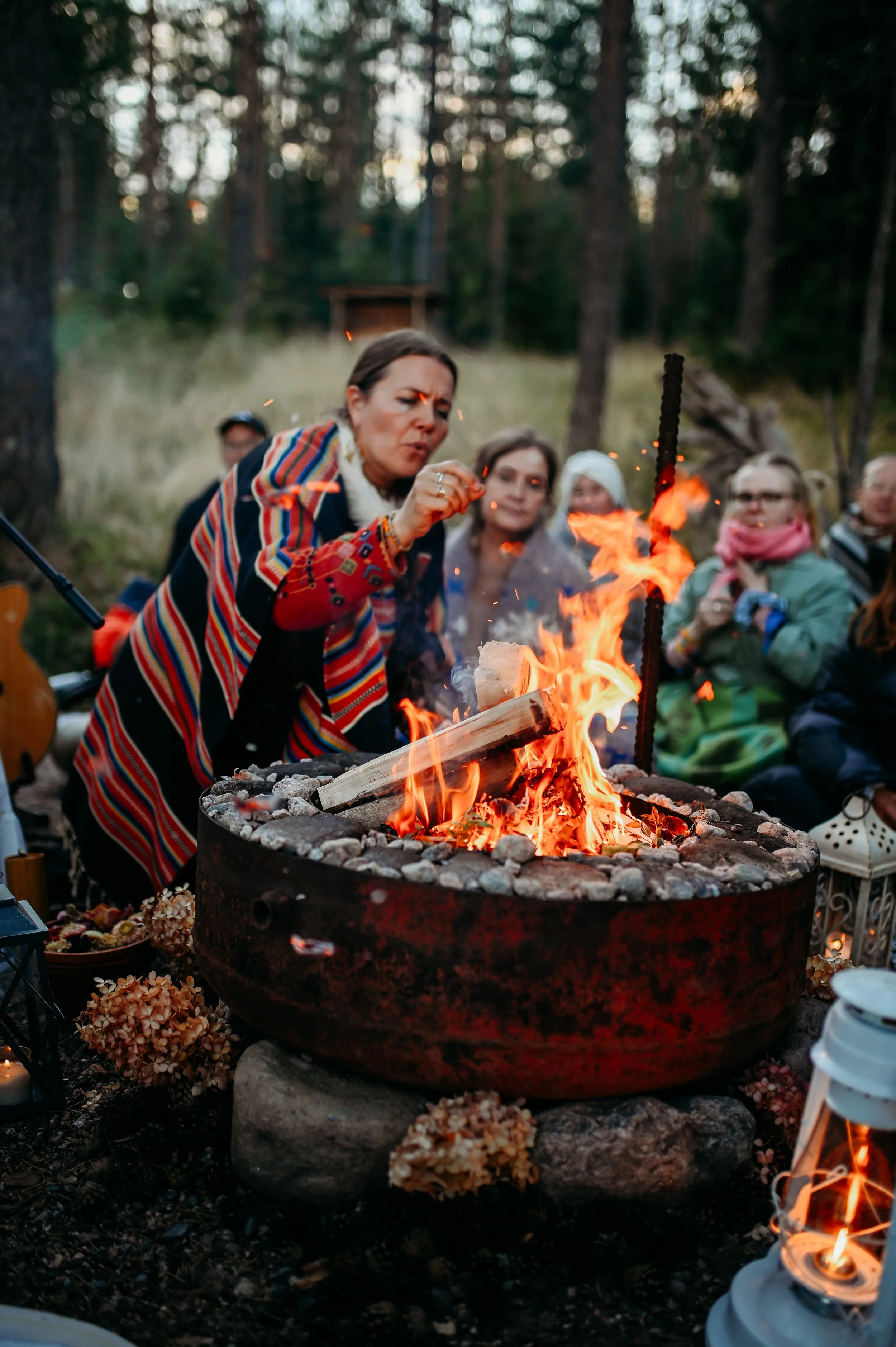 Group of people sitting around a fire pit in a wooded outdoor setting during the evening, with one woman near the fire, wearing a colorful striped shawl, blowing on the fire, while others watch.