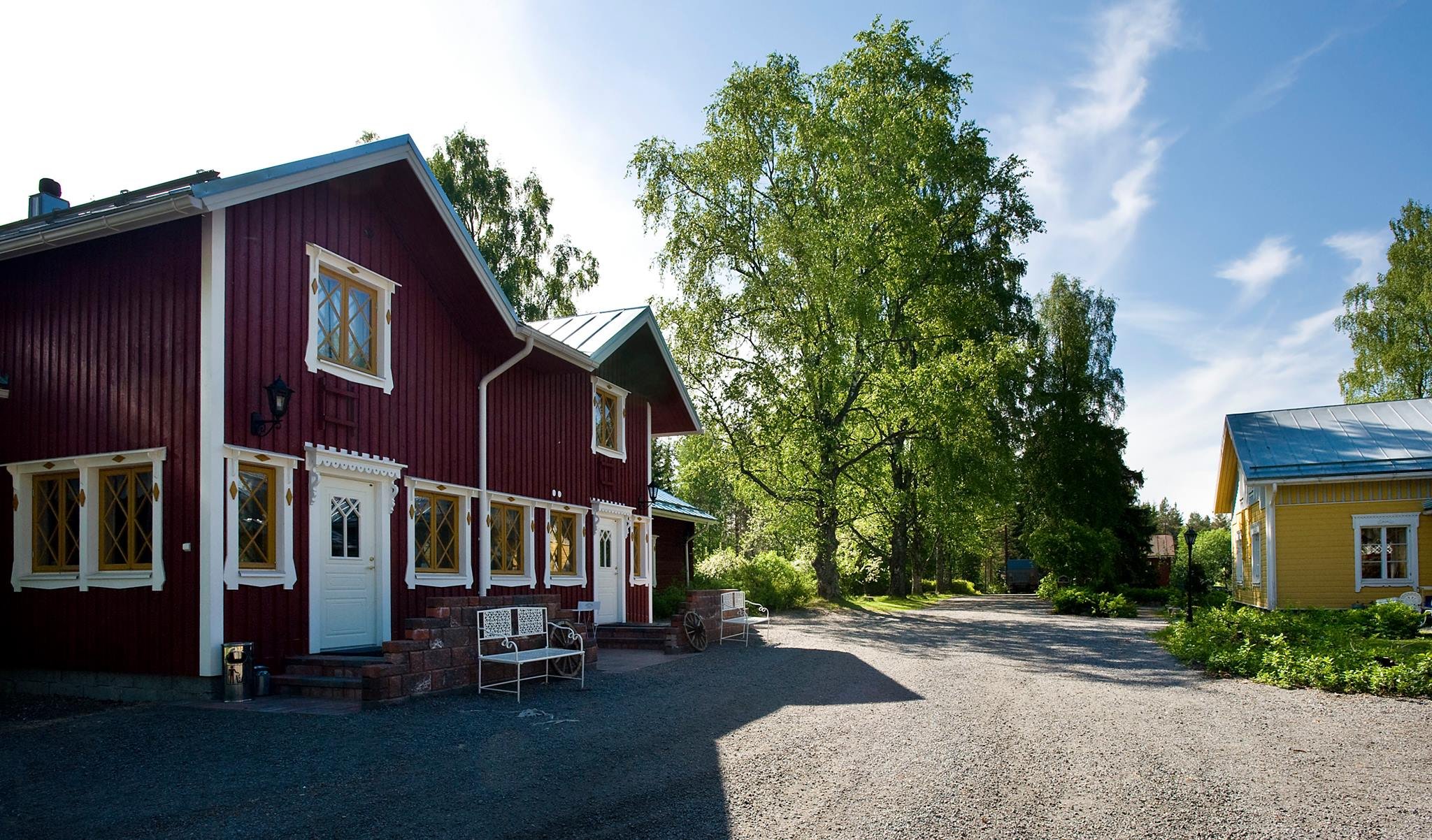 A red two-story house with white trim and diamond-patterned windows, situated outdoors on a sunny day with green trees and clear blue sky in the background.