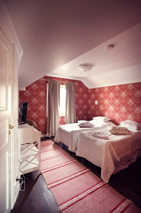 A bedroom with two single beds covered in white bedding, pillows, and a blanket each. The room has patterned red wallpaper, a window with beige curtains, and a pink striped rug on dark wooden floor. There's a small white table with a TV on top, positioned in front of the beds.