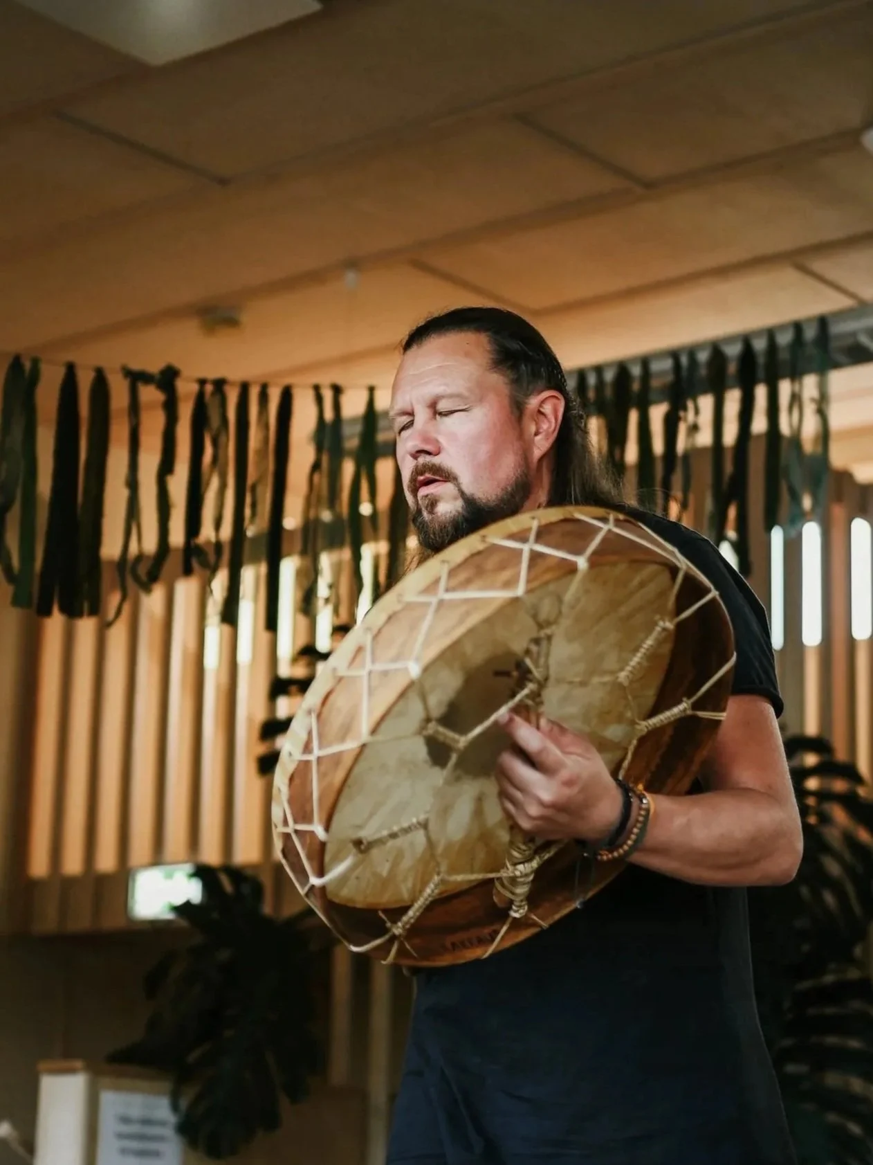 A man with long dark hair and a beard is playing a drum in a room with wooden walls and decorations.