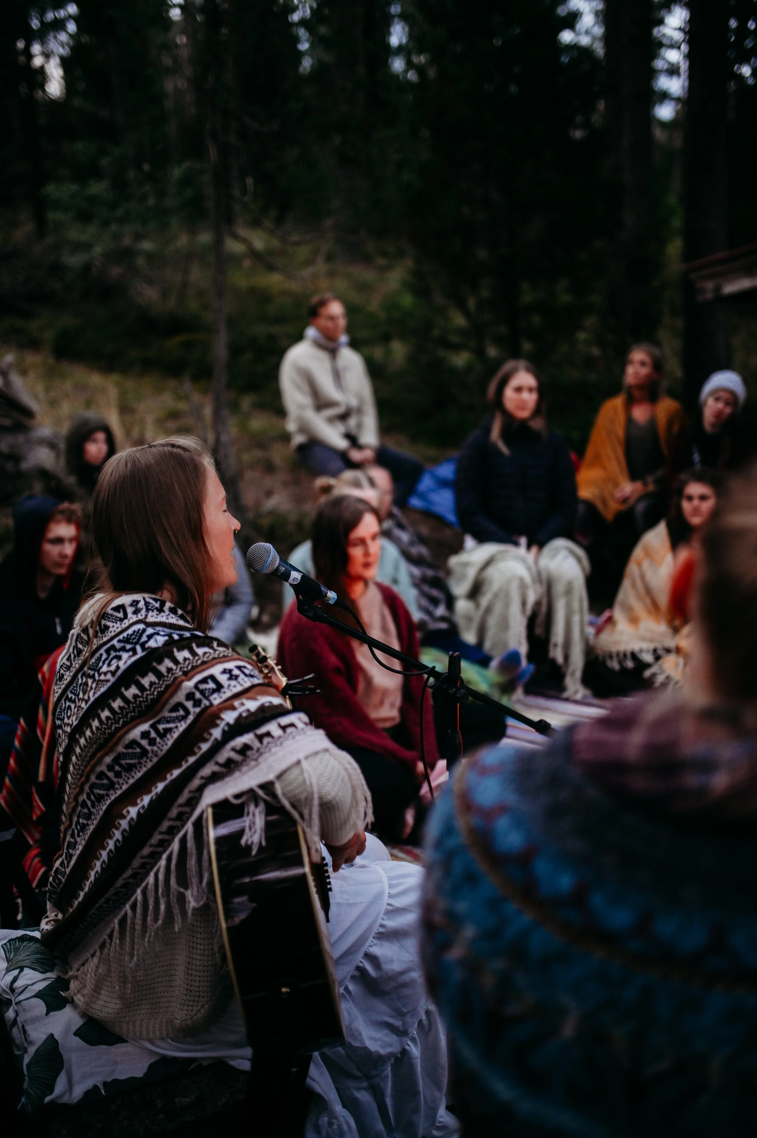A group of people sitting outdoors in a wooded area, listening to a woman speak into a microphone. The scene occurs during twilight or evening.