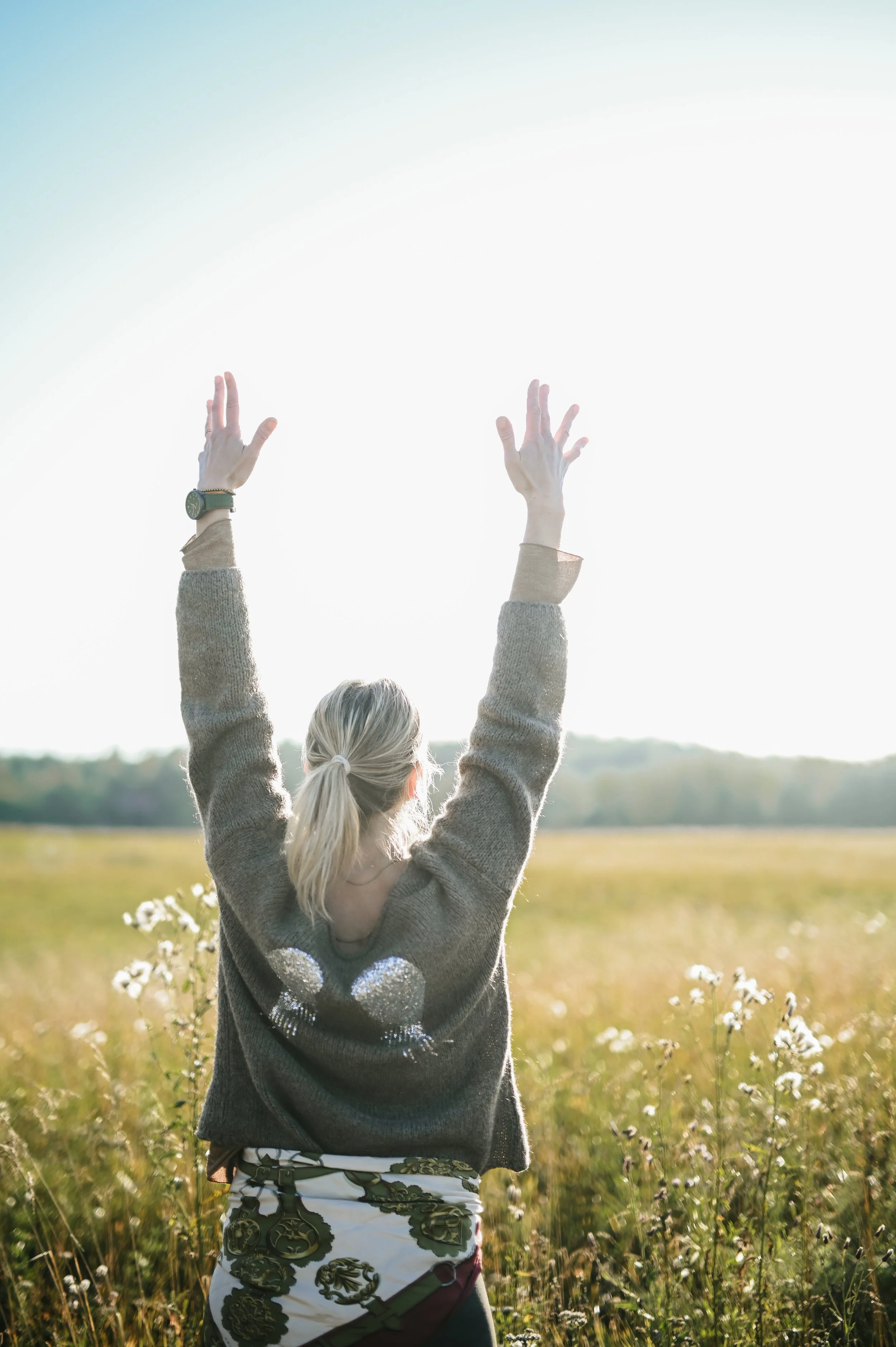 A person with blonde hair tied in a ponytail standing in a field with tall grass and white flowers, raising both hands towards the sky during sunset or sunrise.