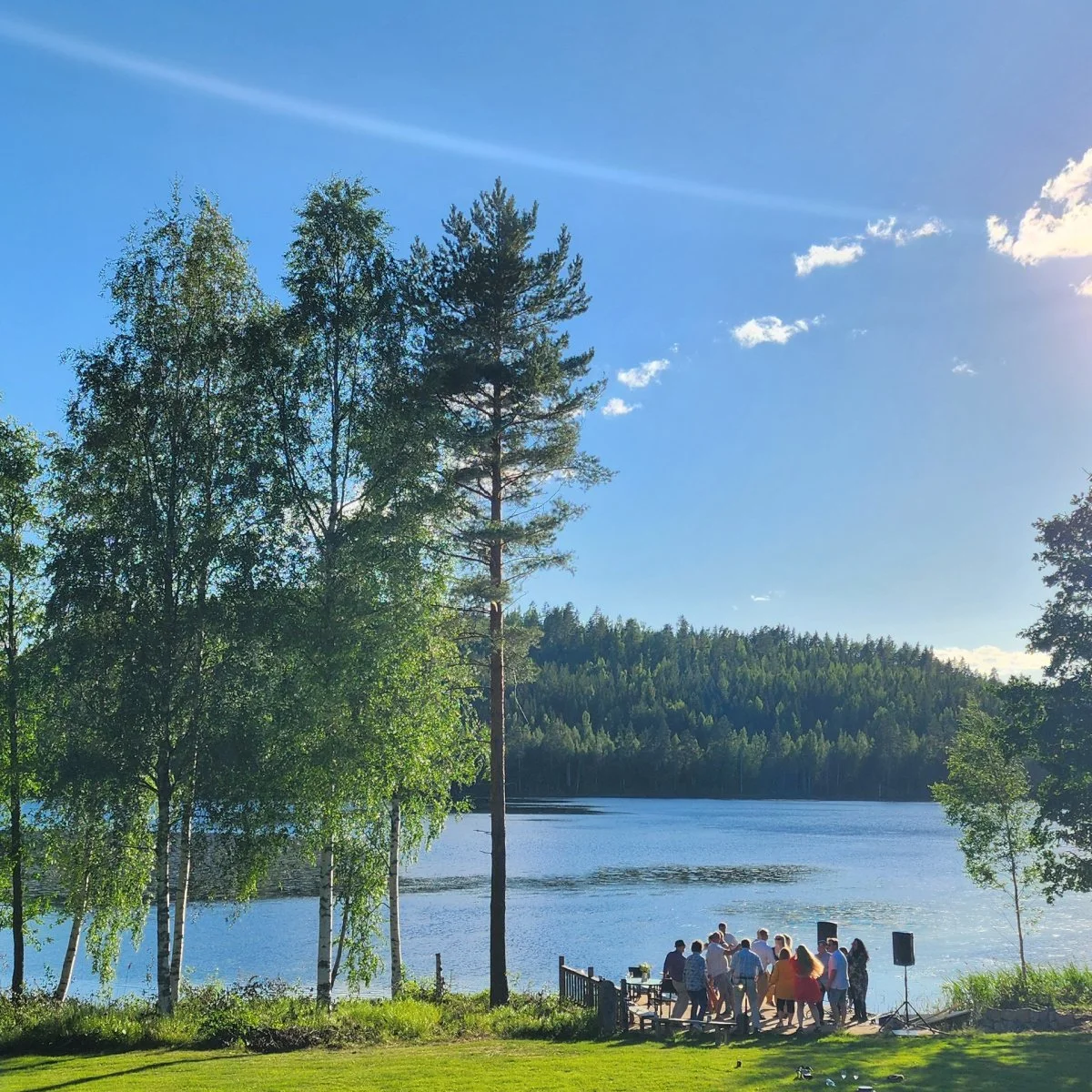 People gathered near a lake with trees and a forest in the background on a sunny day.